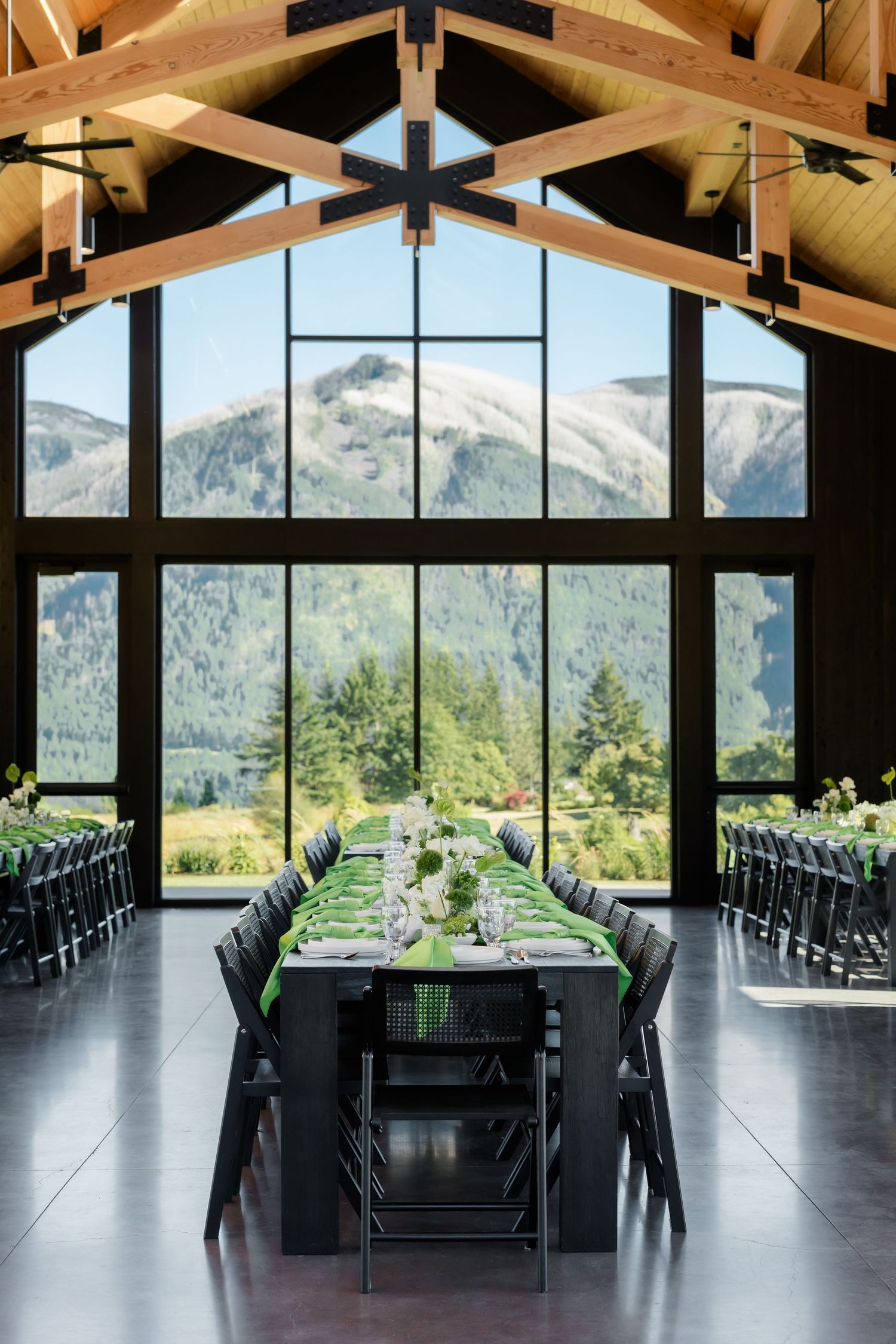 Elegant mountain wedding venue with long black tables, green runners, and floor-to-ceiling windows framing stunning alpine views.