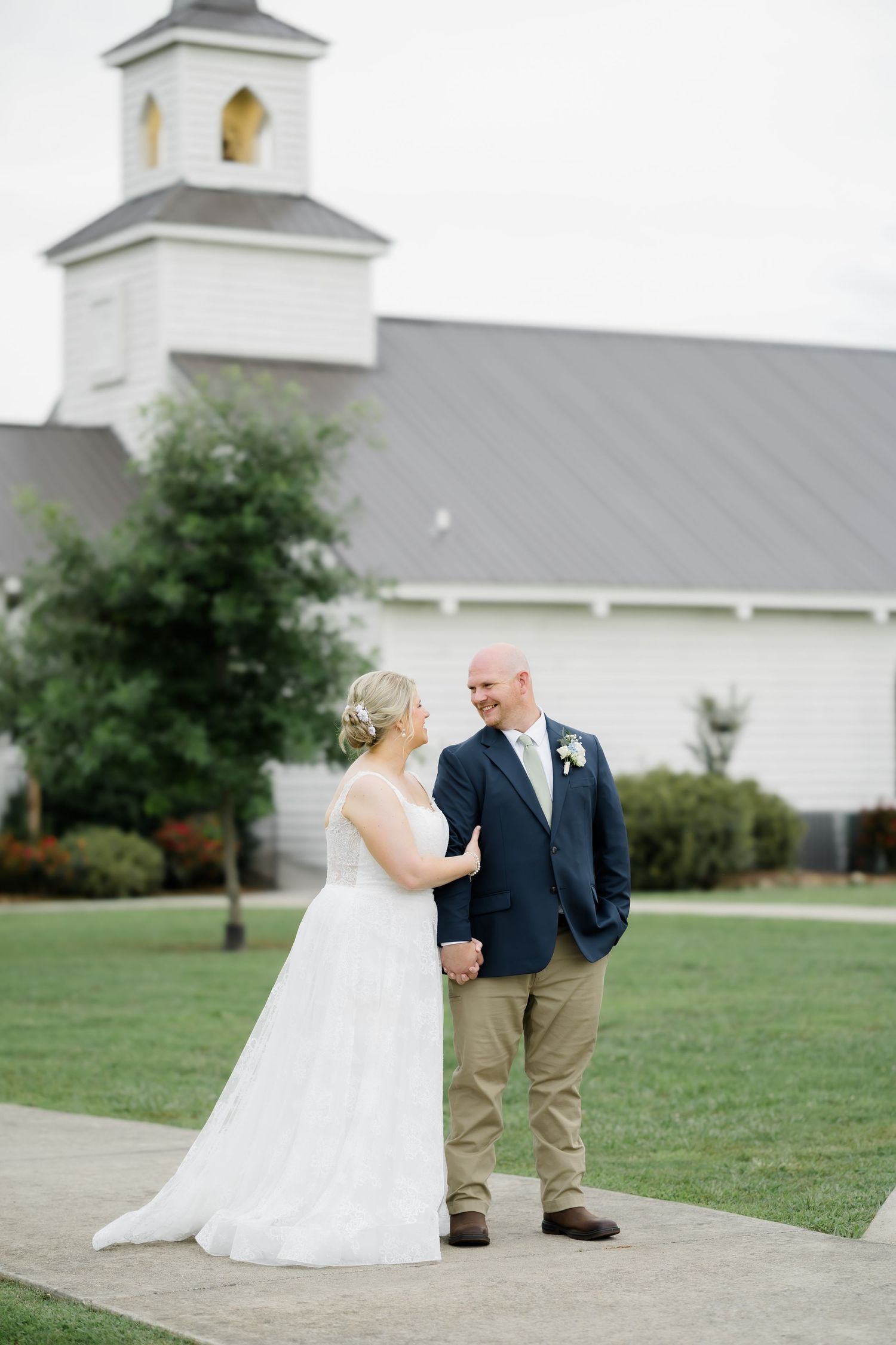Bride and groom hold hands smiling at each other outside a white chapel on their wedding day.