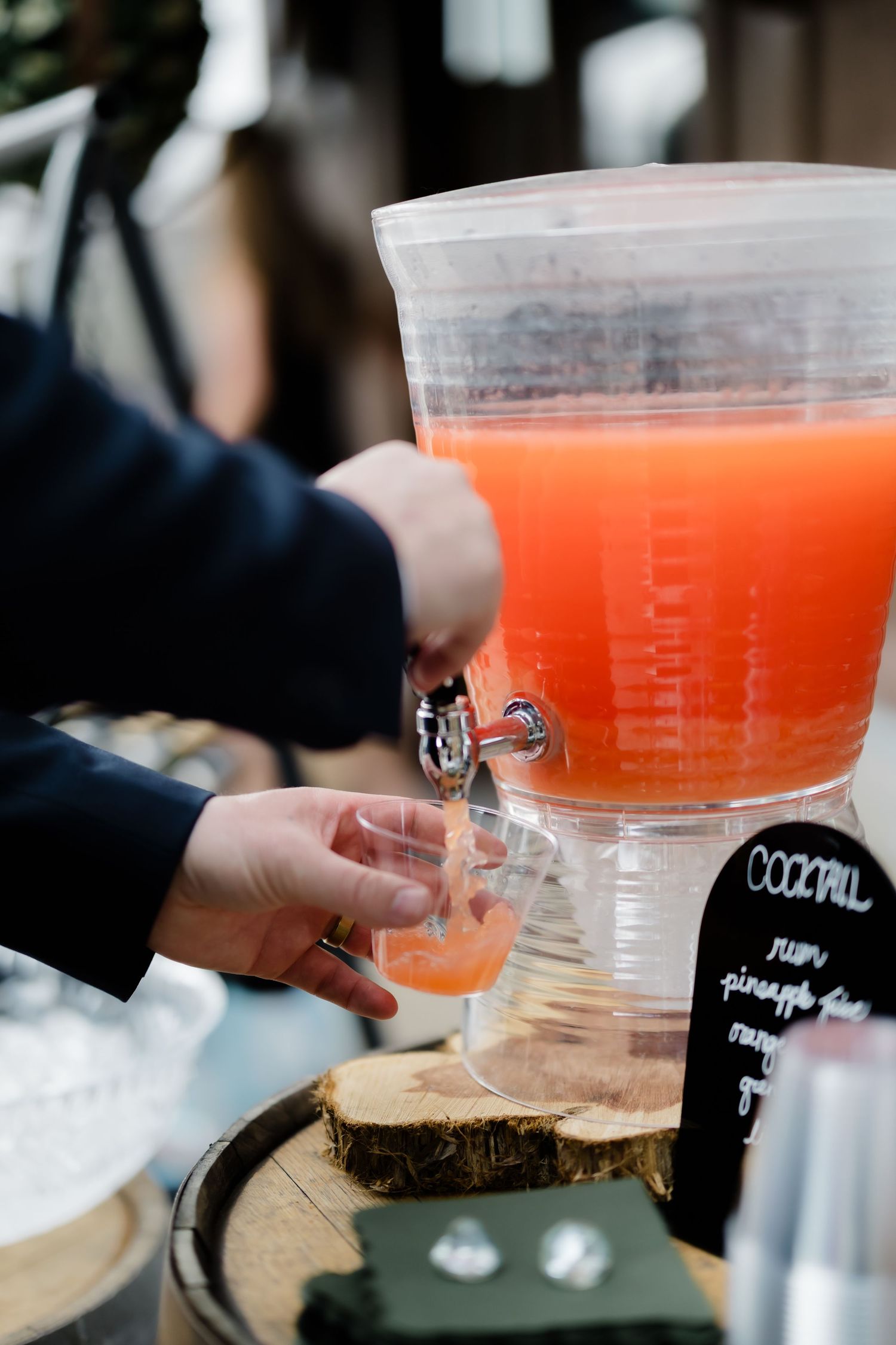 Person pouring orange cocktail punch from large dispenser at outdoor bar station with chalkboard sign.