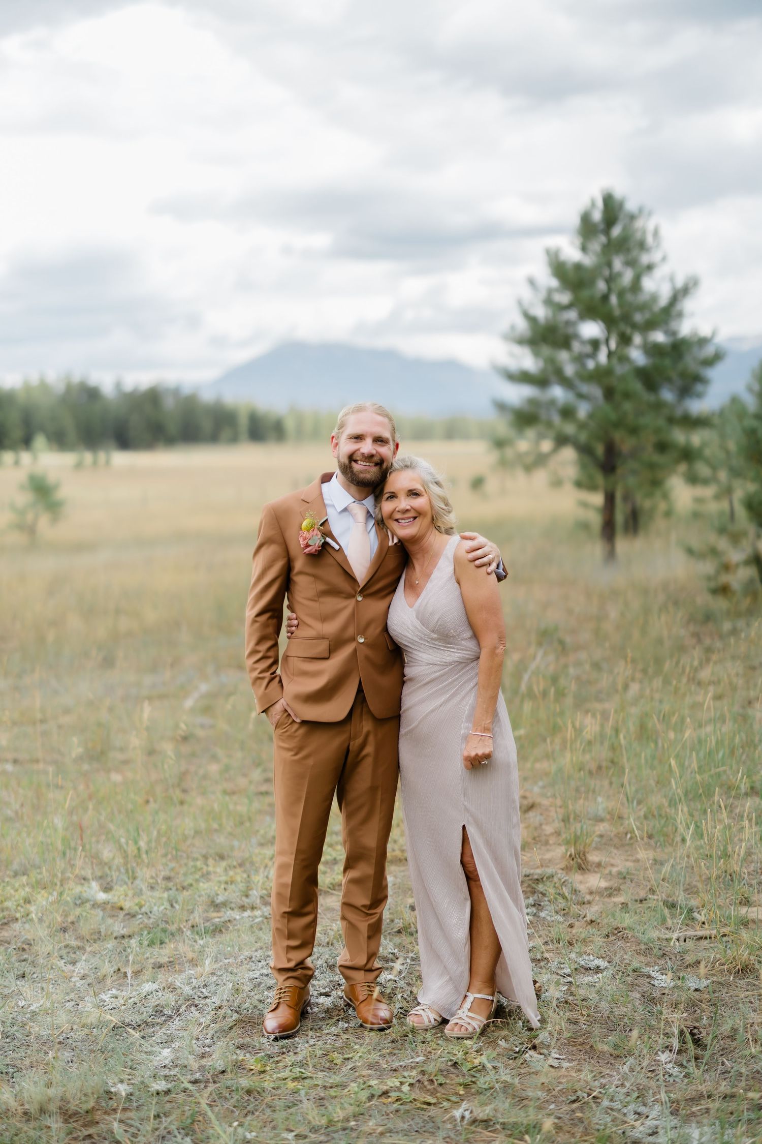 A groom in a brown suit and a woman in a beige gown pose together in a mountain meadow at an outdoor wedding.