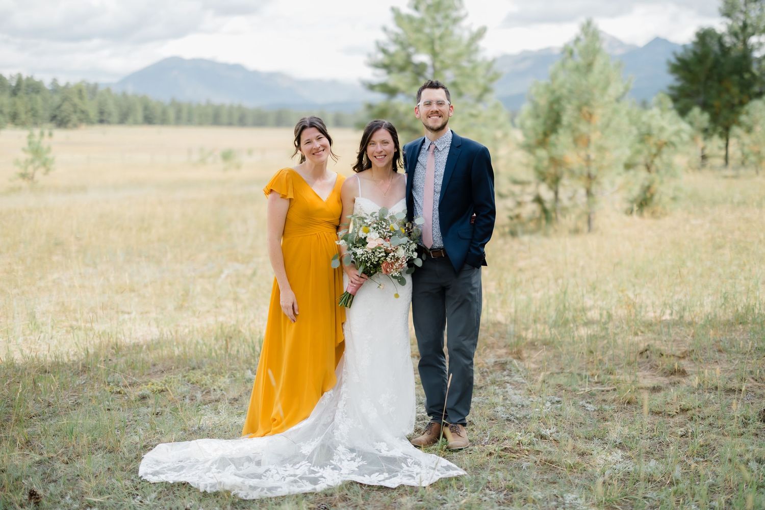 Bride in white gown and bridesmaid in yellow dress pose with groom in navy suit in a mountain meadow.