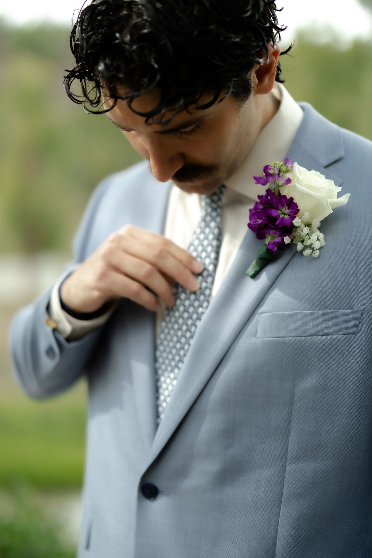Groom in light blue suit adjusting tie with purple and white boutonniere on lapel at wedding.