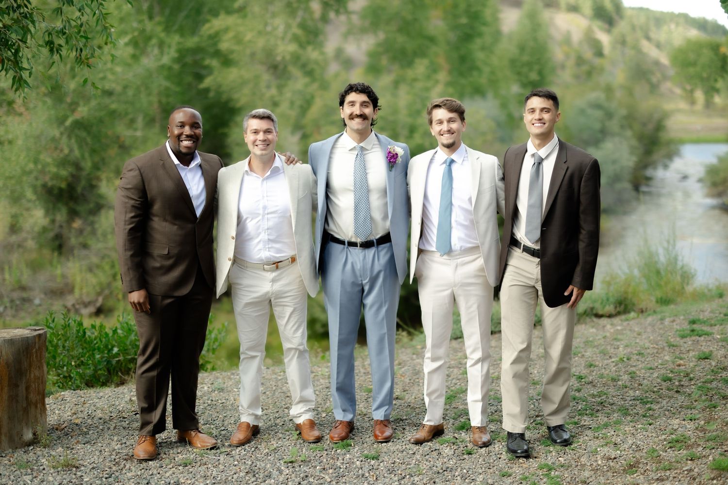 Five groomsmen in suits and ties posing together outdoors near a river at a wedding celebration.