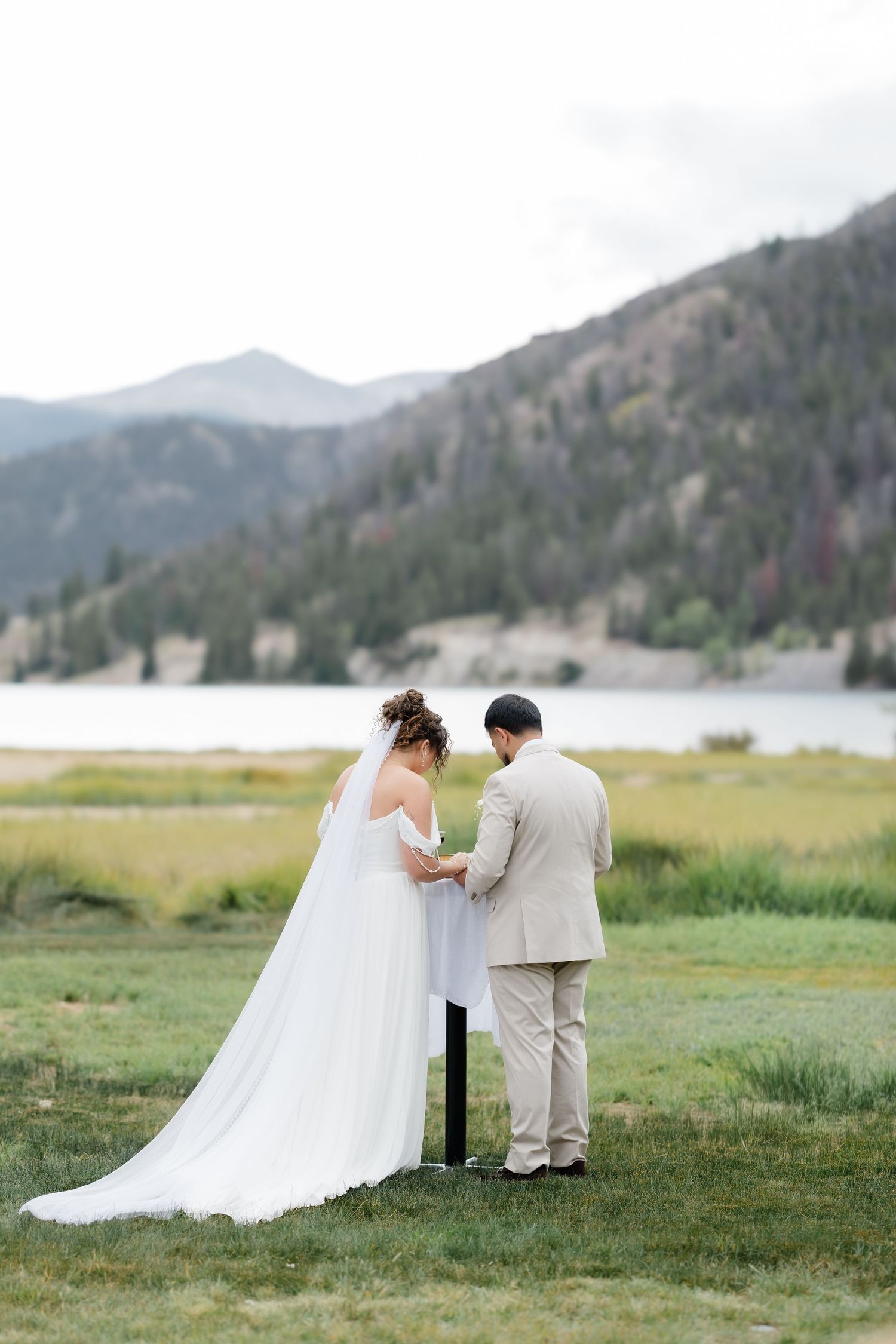 Bride and groom hold hands at outdoor mountain lake wedding ceremony on lush green grass.