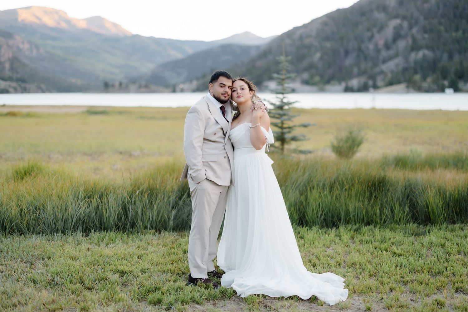 A bride and groom pose together in a white gown and tan suit with mountain lake scenery behind them.