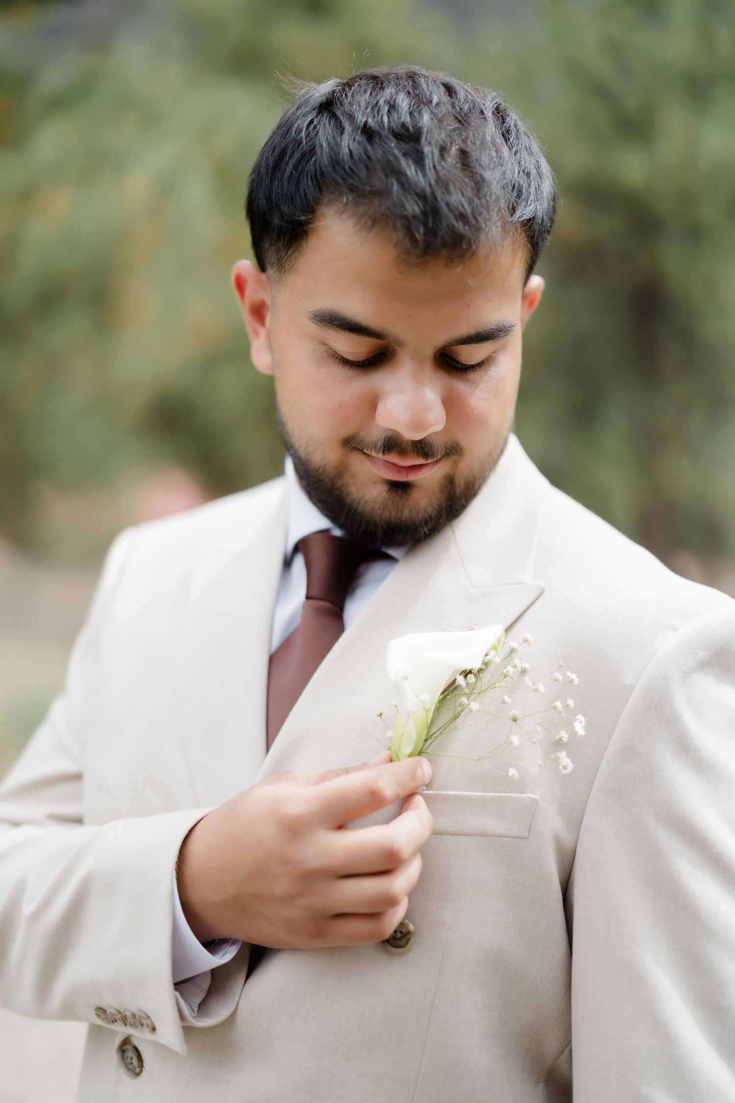 Groom in beige suit adjusting white rose boutonniere, wearing mauve tie, with greenery background.