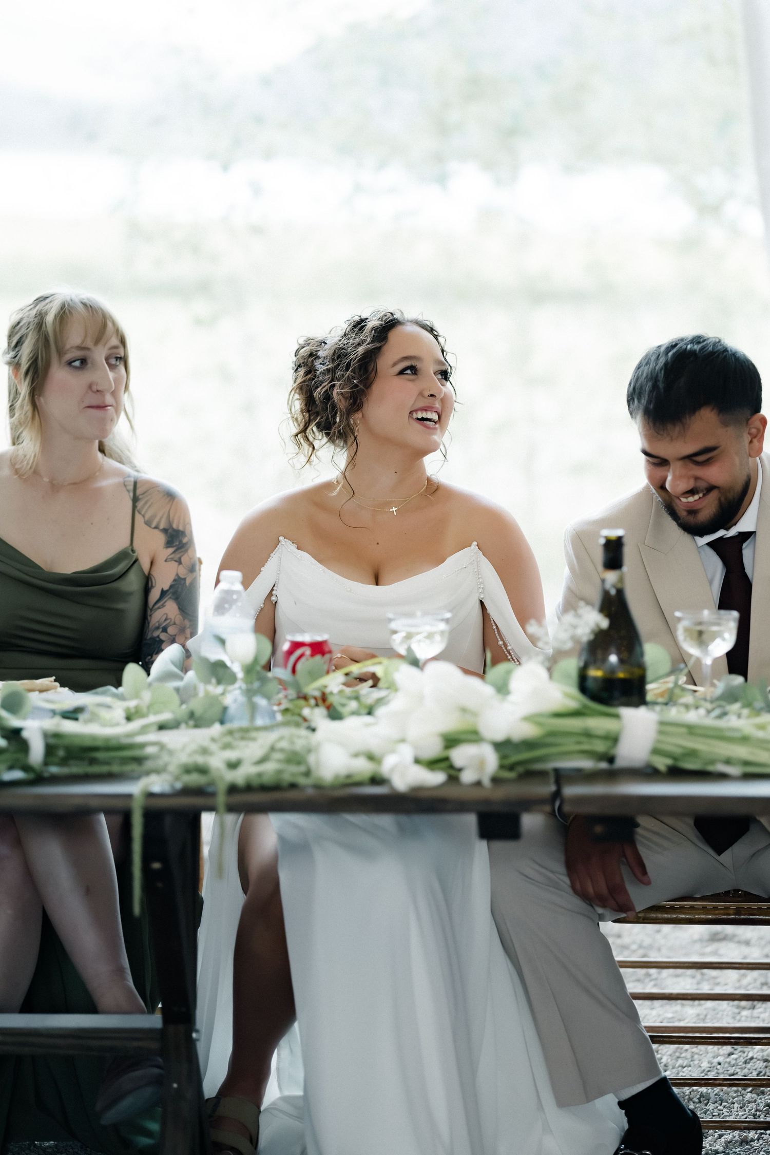 Radiant bride laughs joyfully at head table with bridesmaids and groom at wedding reception.