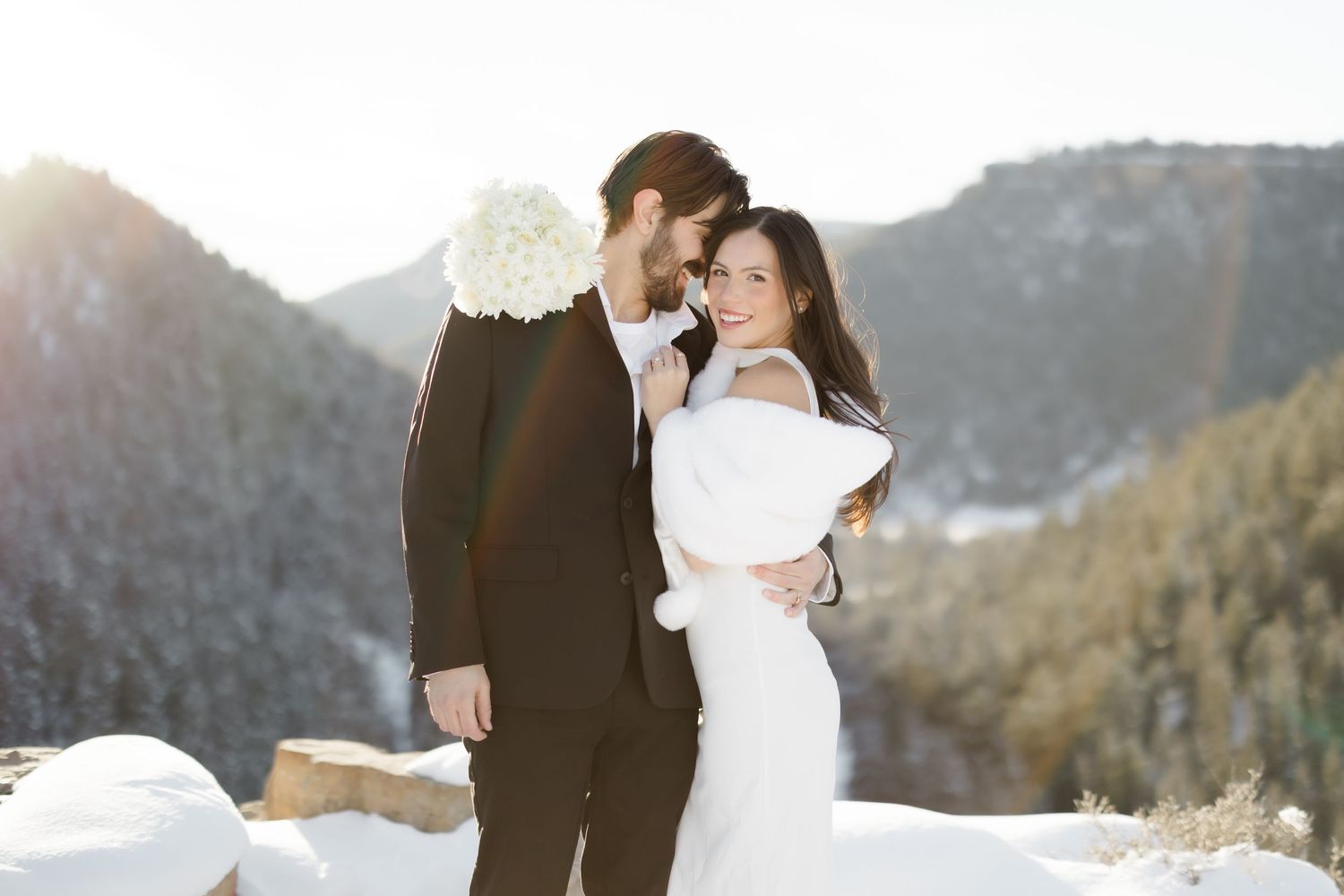 Bride and groom embrace in snowy mountain landscape, bride holding white bouquet in elegant winter wedding attire.