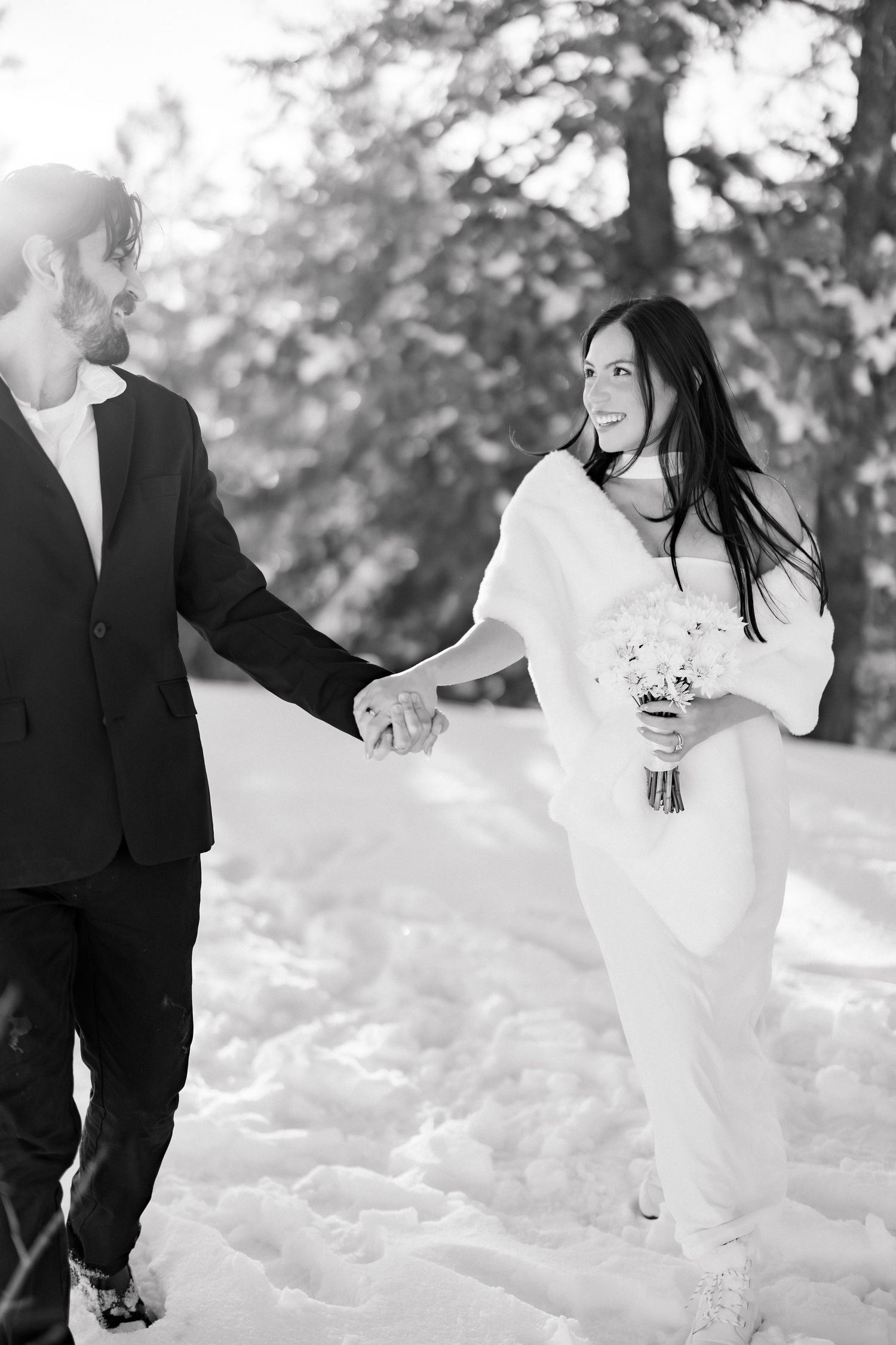 Black and white photo of bride and groom holding hands walking through snow among winter trees.