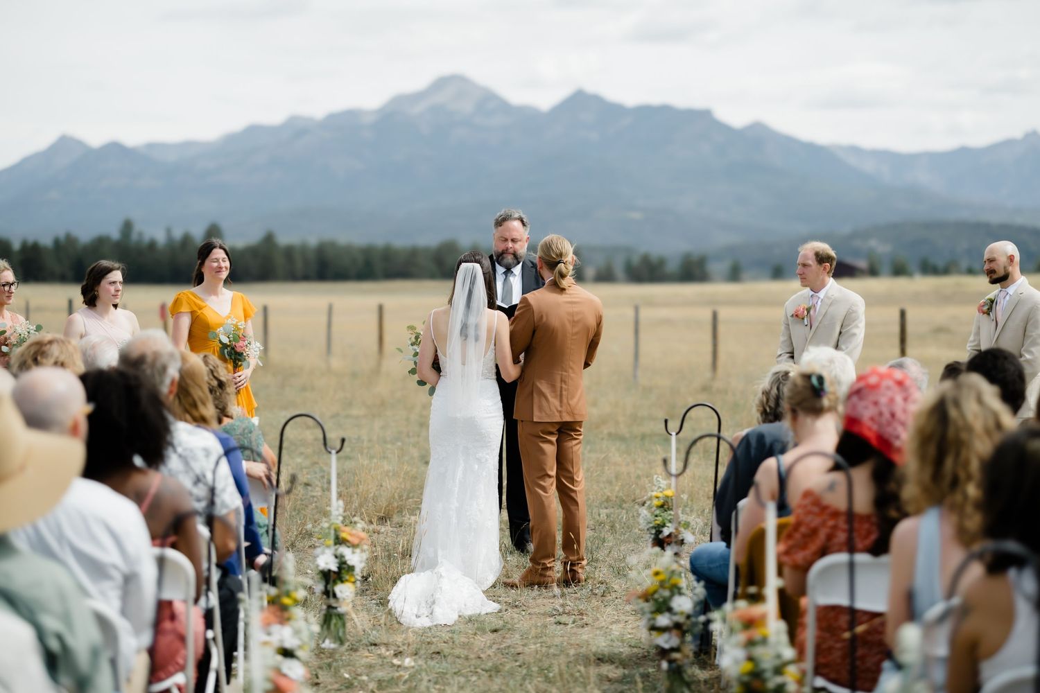 Outdoor wedding ceremony in a mountain meadow with bride and groom at altar, guests seated, and snow-capped peaks in background.