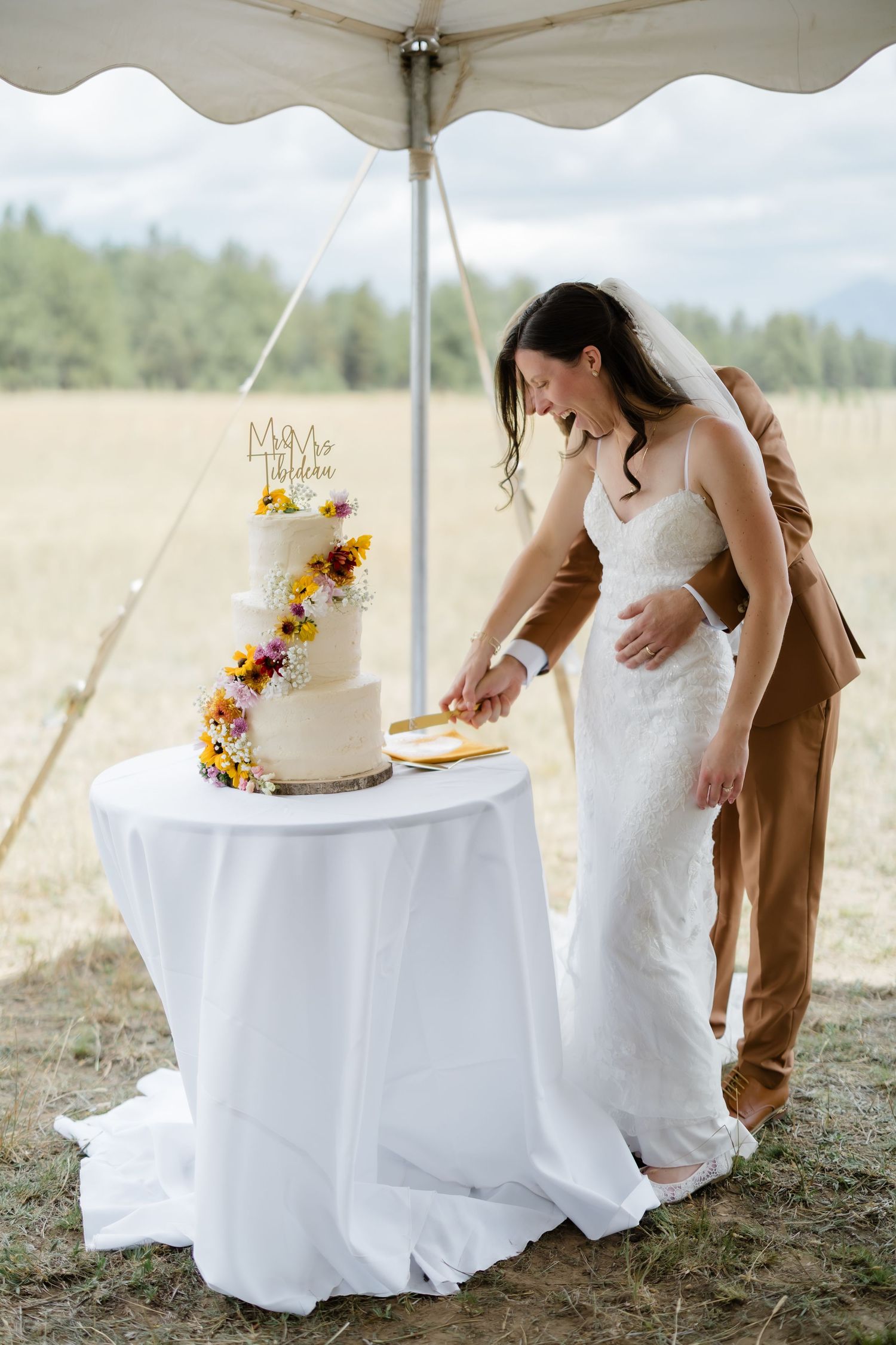 Bride and groom cutting a floral three-tier wedding cake on a white draped table under an outdoor tent.