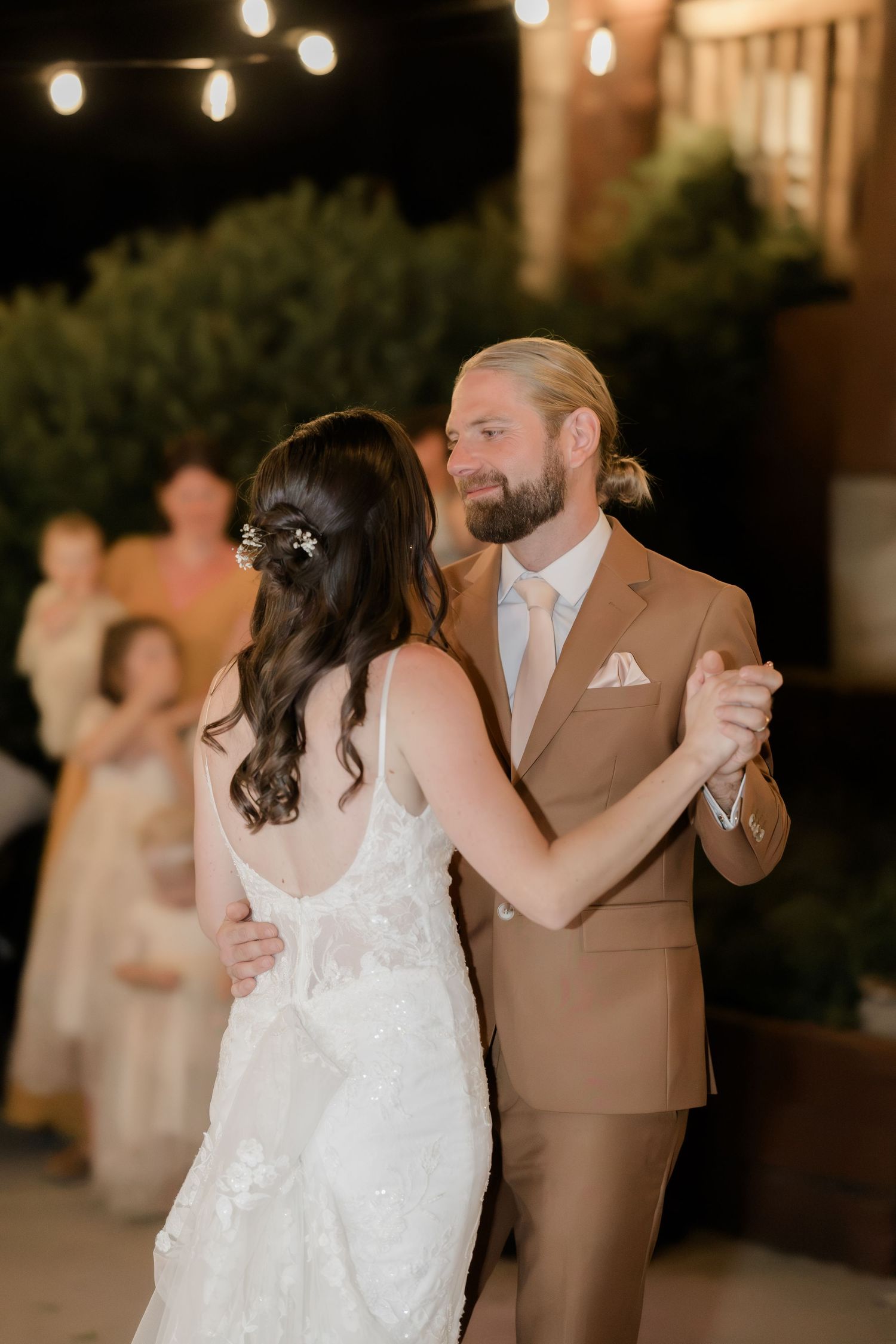 Bride and groom share their first dance outdoors at night, with string lights and guests visible in the background.