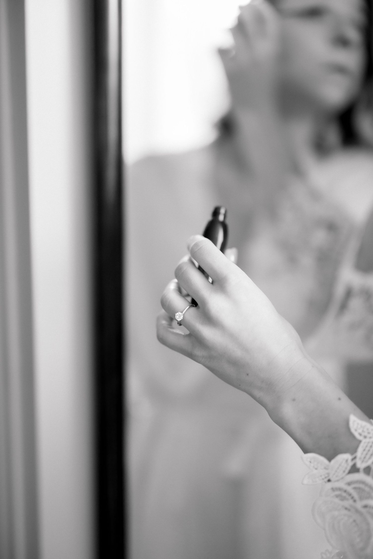 Bride applying perfume while wearing an engagement ring, reflected in a mirror, captured in black and white.