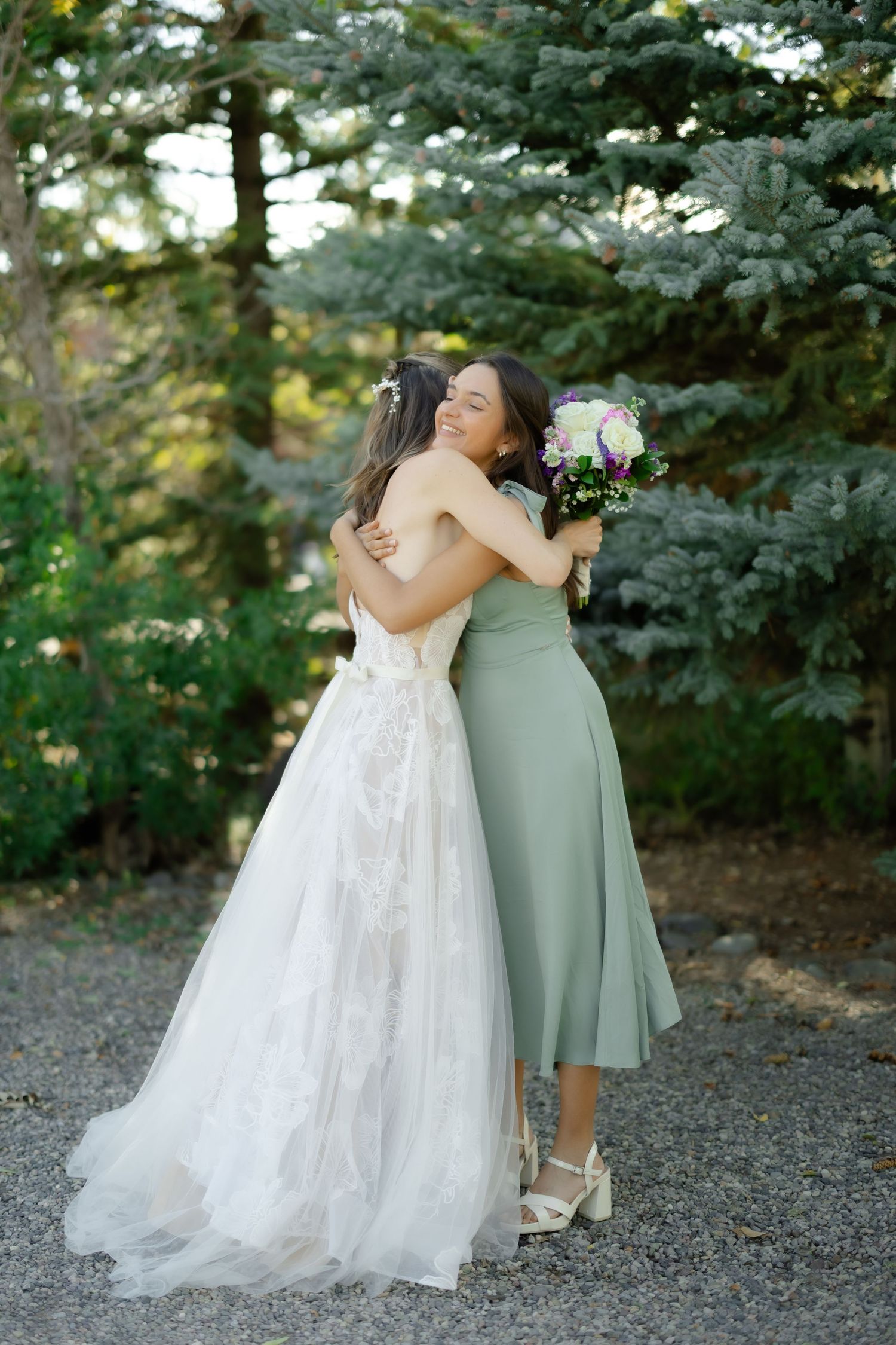 A bride in a white lace gown hugs a bridesmaid in a sage green dress, holding a bouquet, surrounded by evergreen trees.