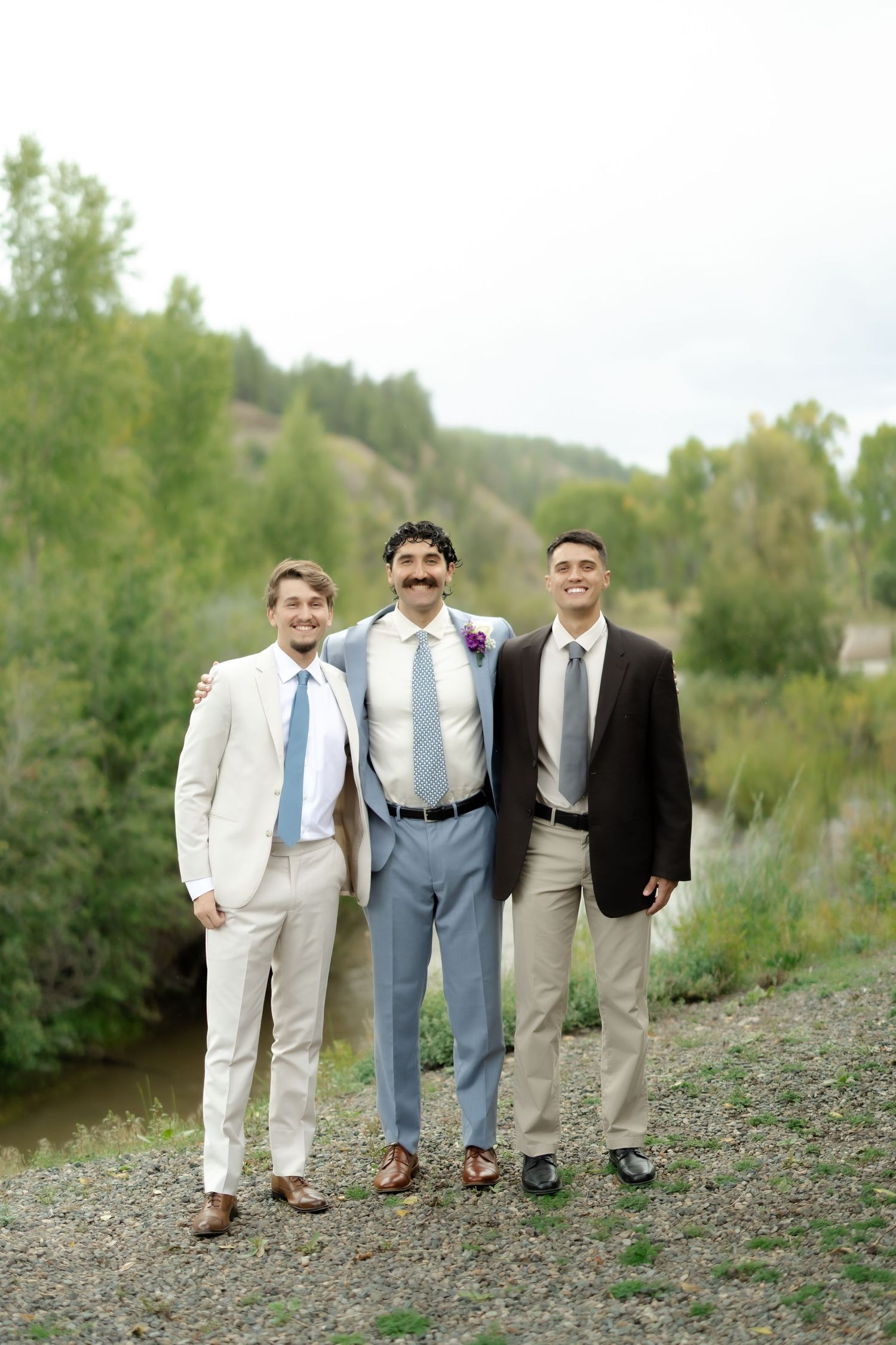 Three men in suits smiling outdoors near a river at a wedding, with lush green trees in the background.