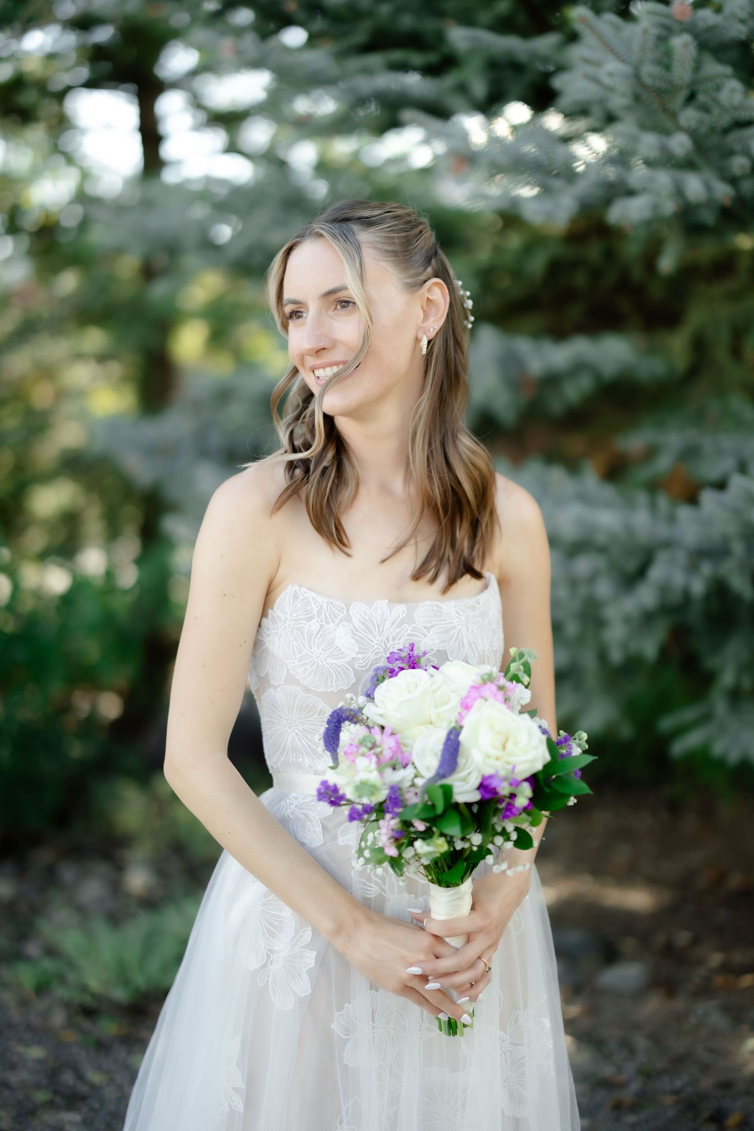 Smiling bride in strapless lace wedding gown holding purple and white bouquet outdoors among evergreen trees.