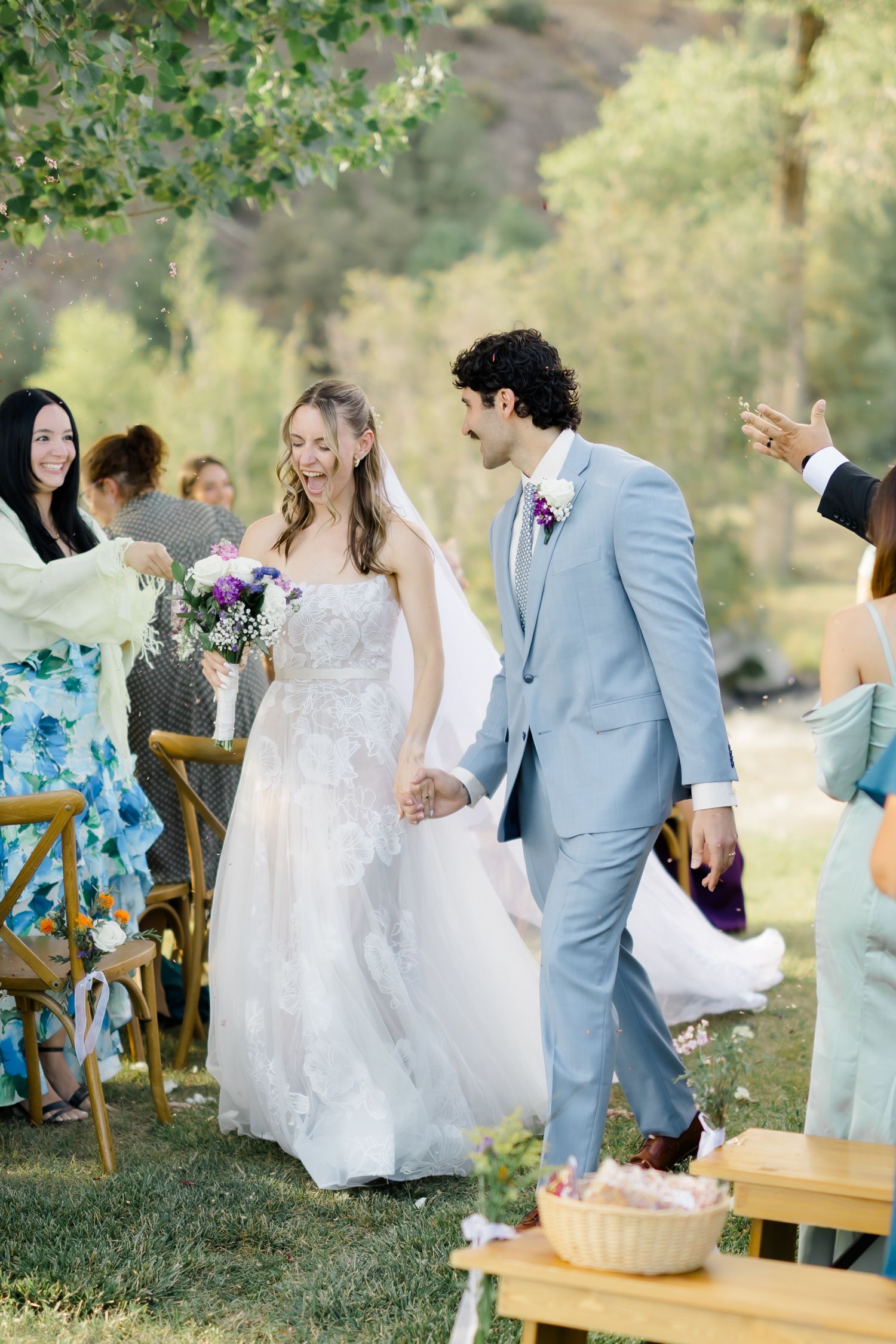Bride and groom walk down outdoor aisle hand in hand, smiling, surrounded by guests at a sunny garden wedding.