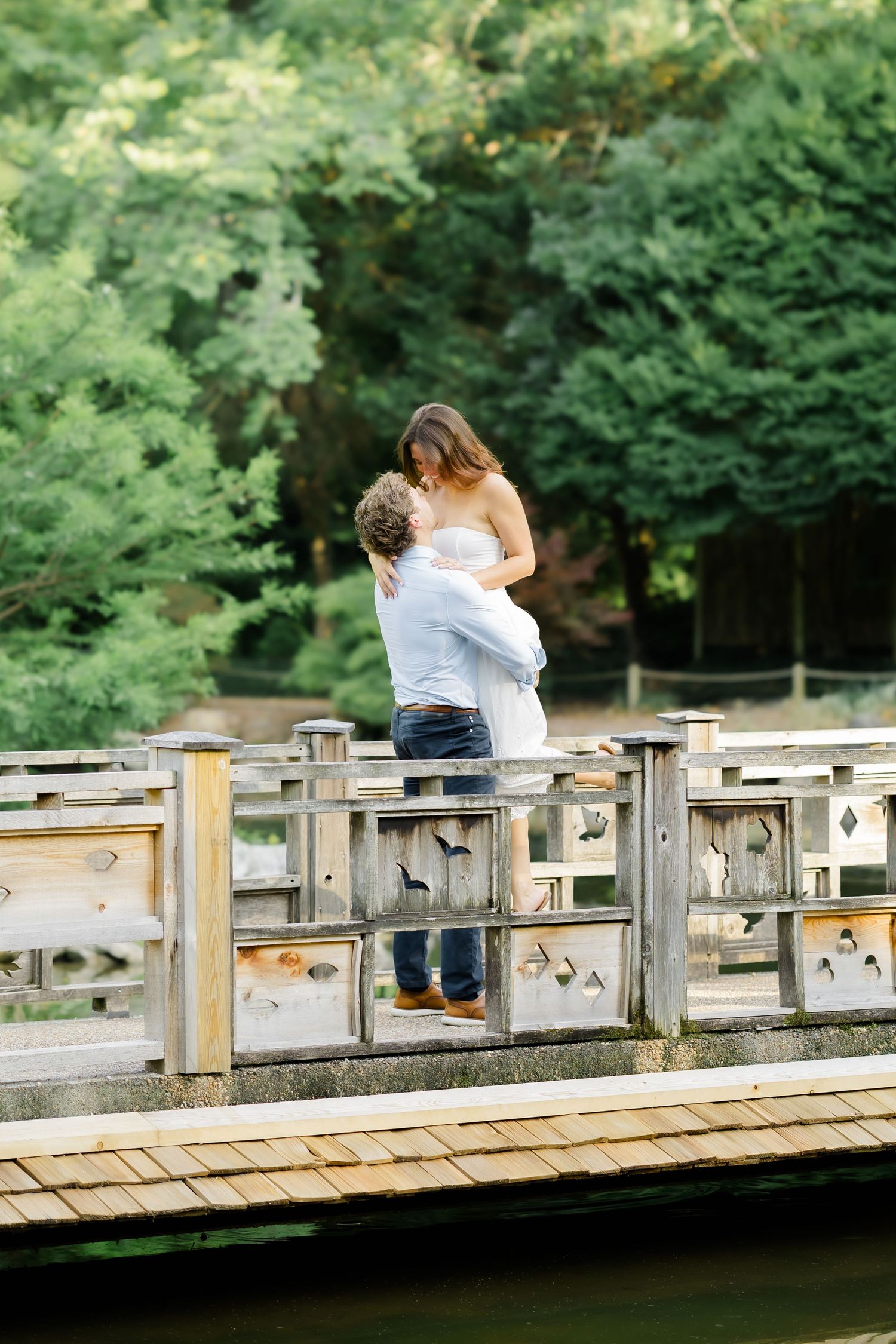 Couple embracing on a wooden bridge surrounded by lush green trees in a romantic outdoor setting.