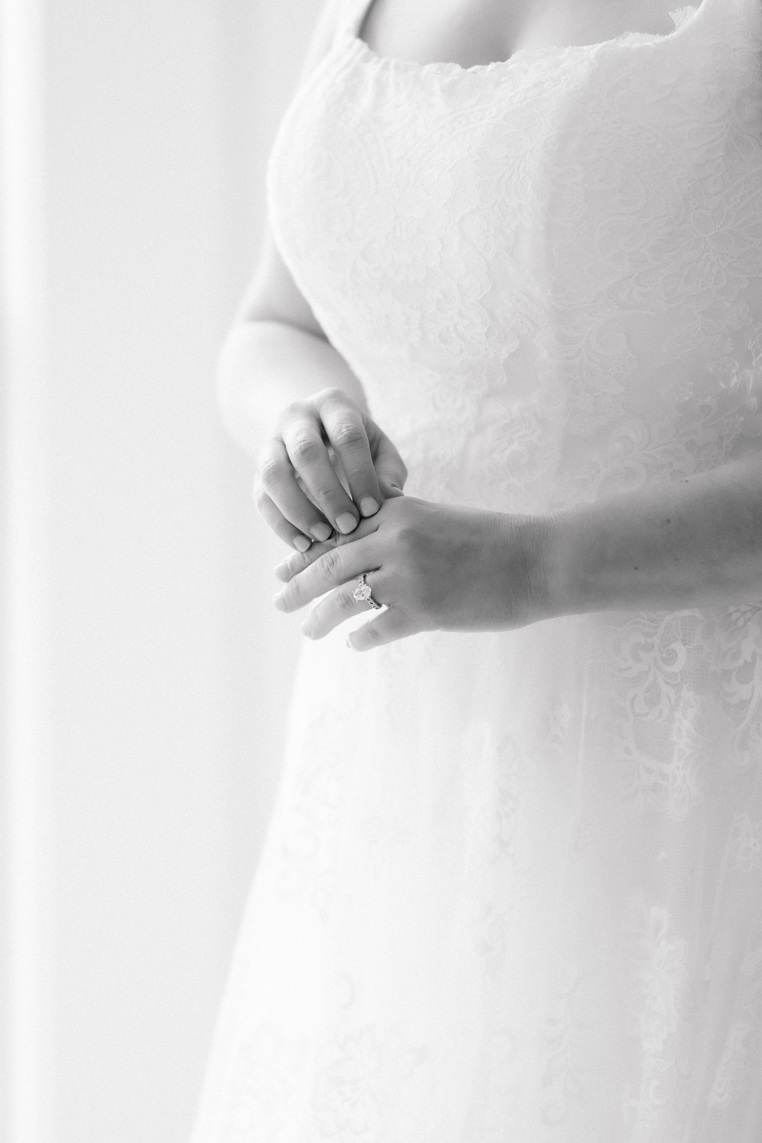 Black and white photo of a bride in a lace wedding dress clasping her hands showing her engagement ring.