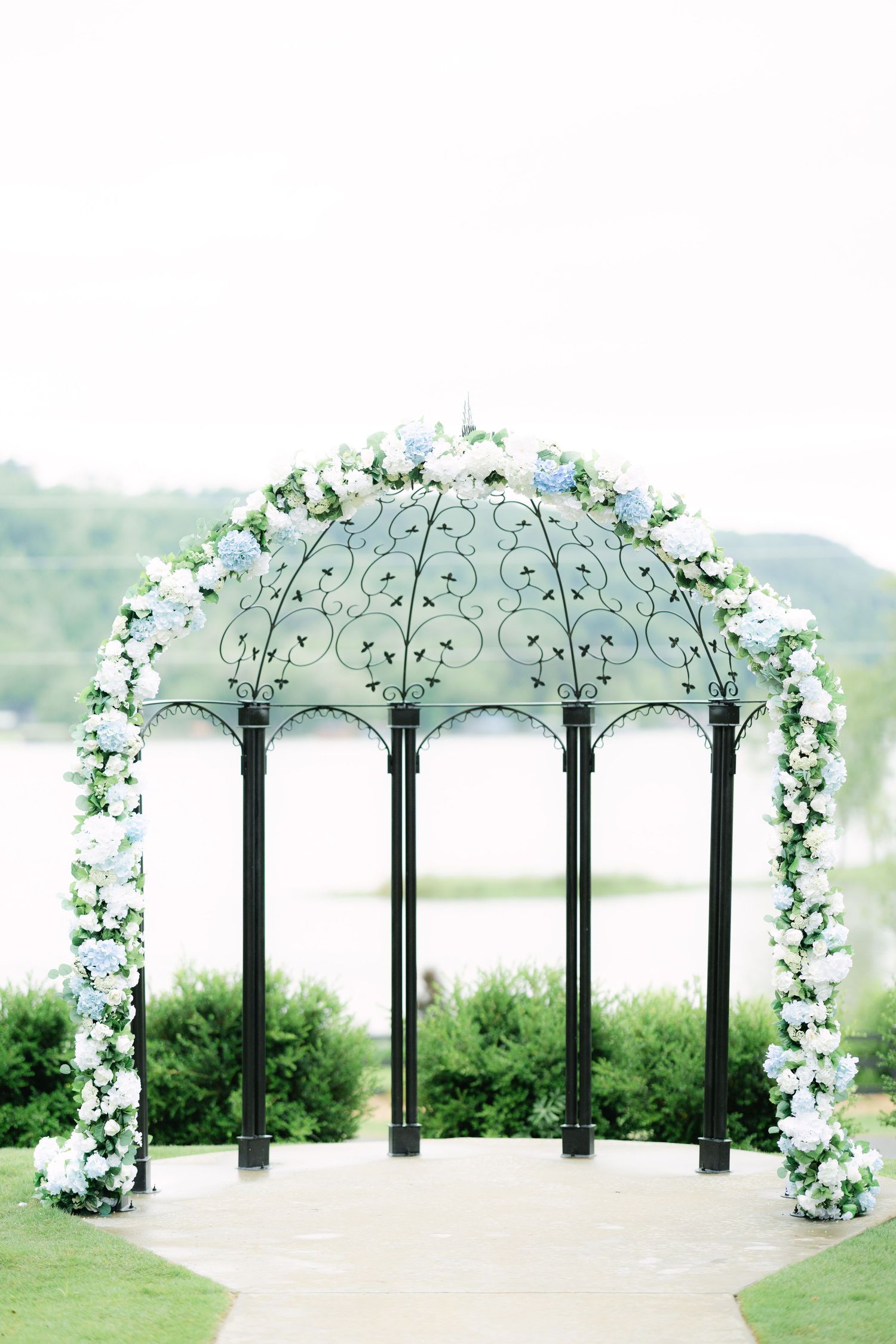 A white floral wedding arch with black metal columns stands elegantly on a grassy hillside overlooking scenic views.