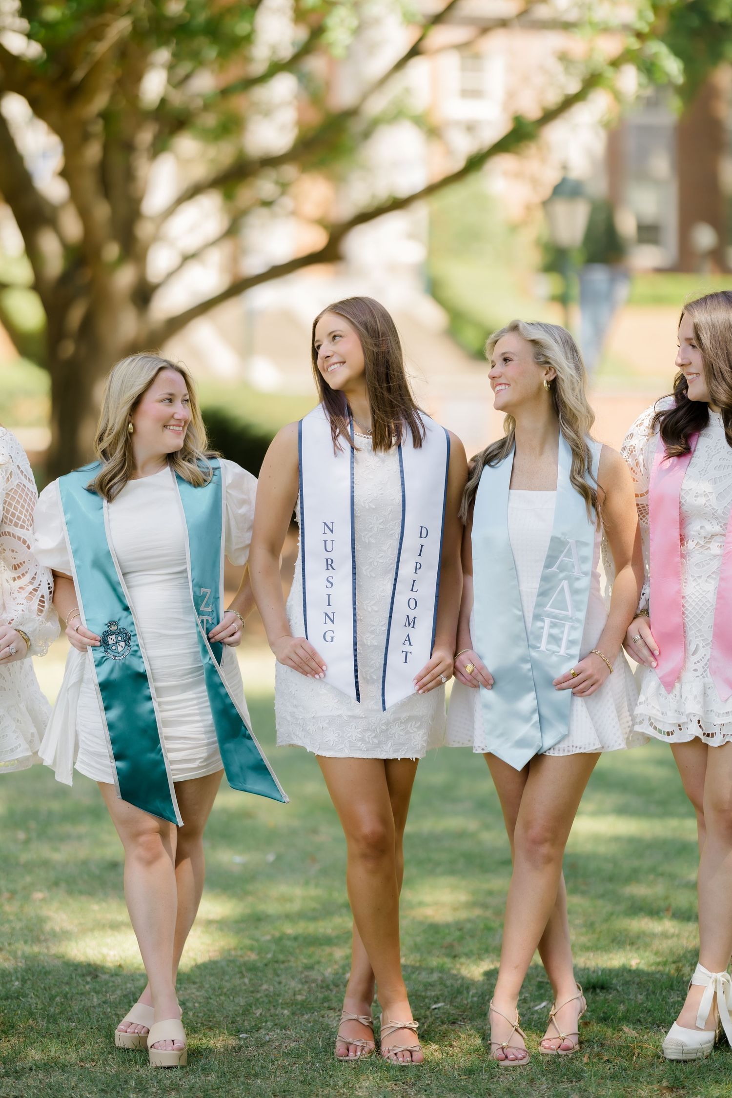 Four young women in white dresses wearing graduation sashes walk outdoors on a college campus celebrating their graduation.