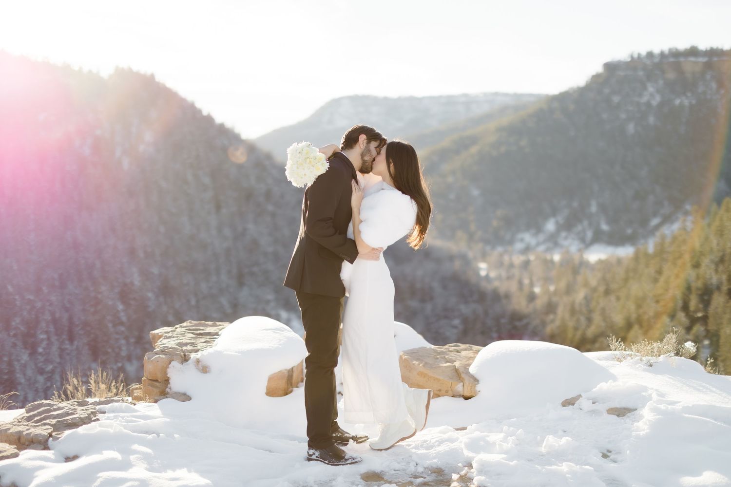 Couple sharing a romantic kiss in snowy mountain landscape during winter wedding elopement with golden sunlight.