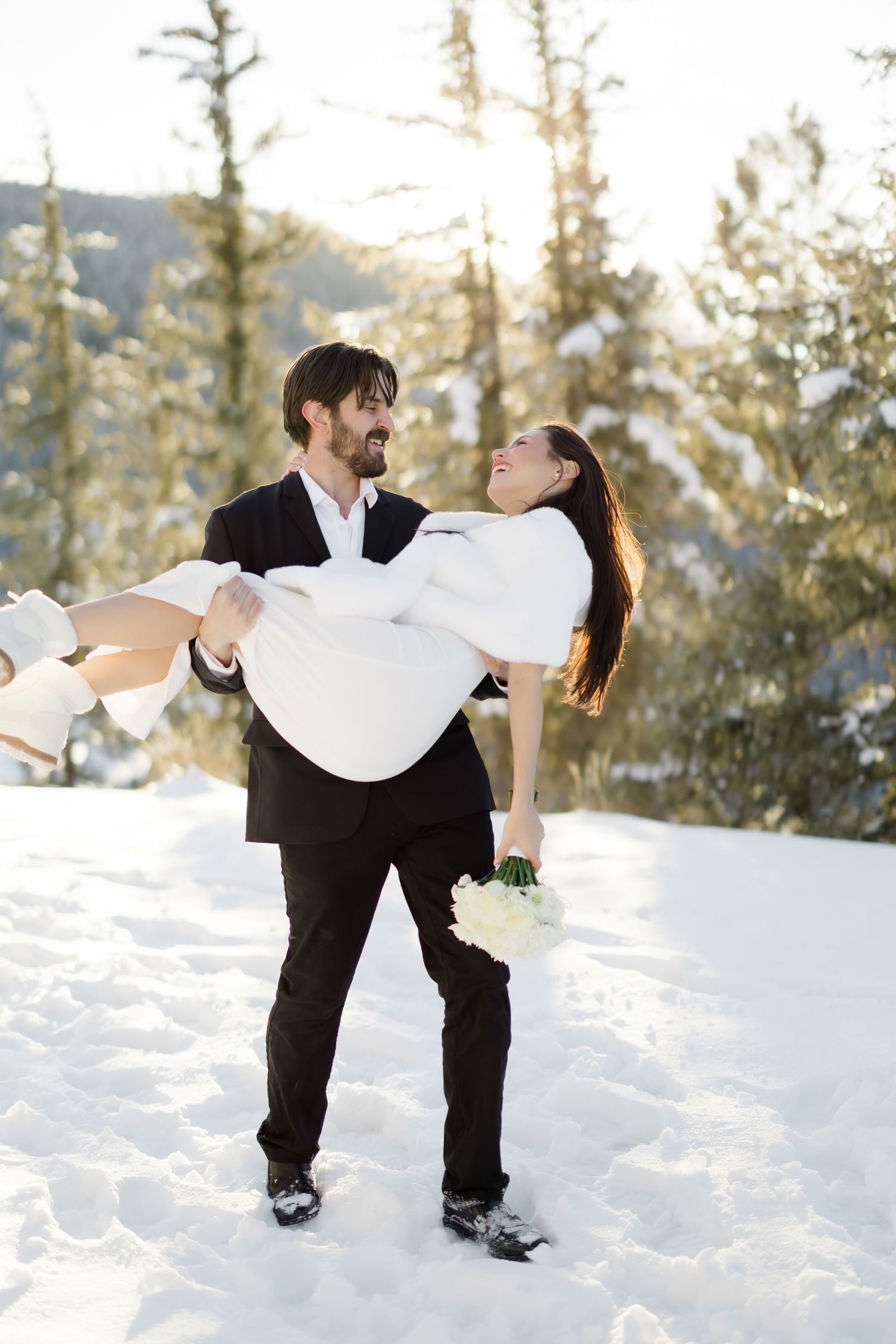 Groom carries laughing bride in his arms through a snowy mountain landscape on wedding day.