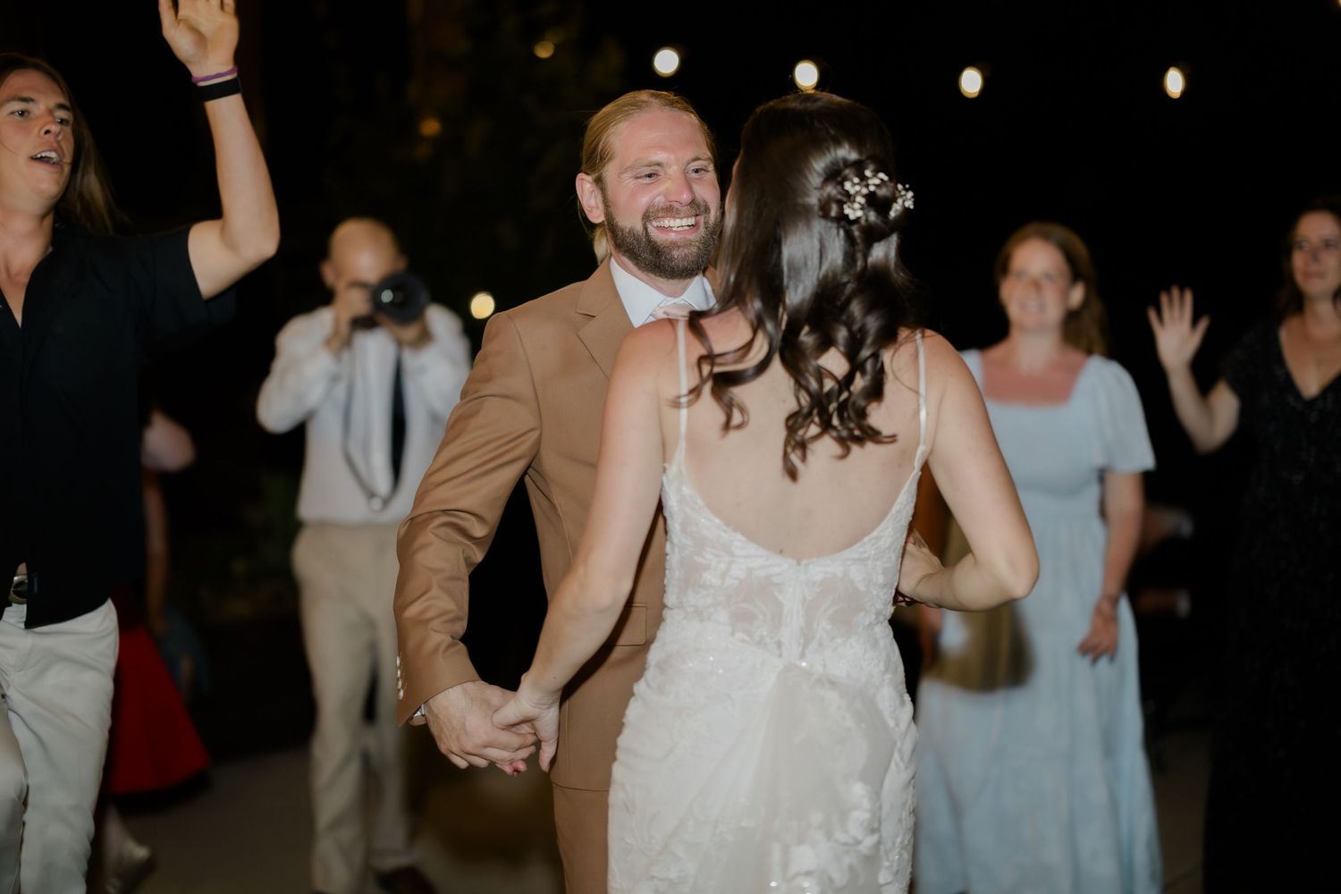 Smiling groom in tan suit holds bride in white lace dress during first dance at outdoor wedding reception.