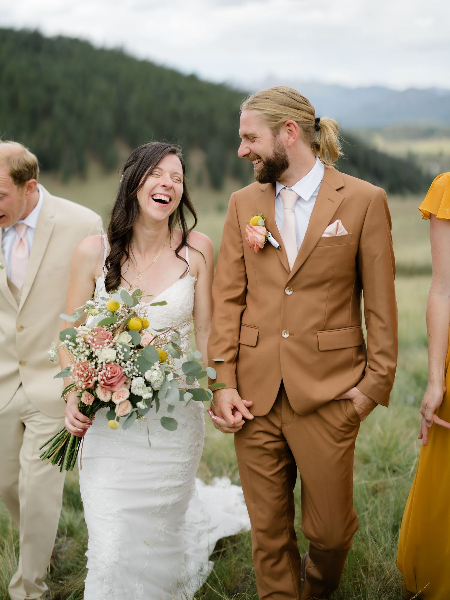 Laughing bride in white gown holds floral bouquet beside groom in tan suit, walking outdoors with wedding party in mountain meadow.