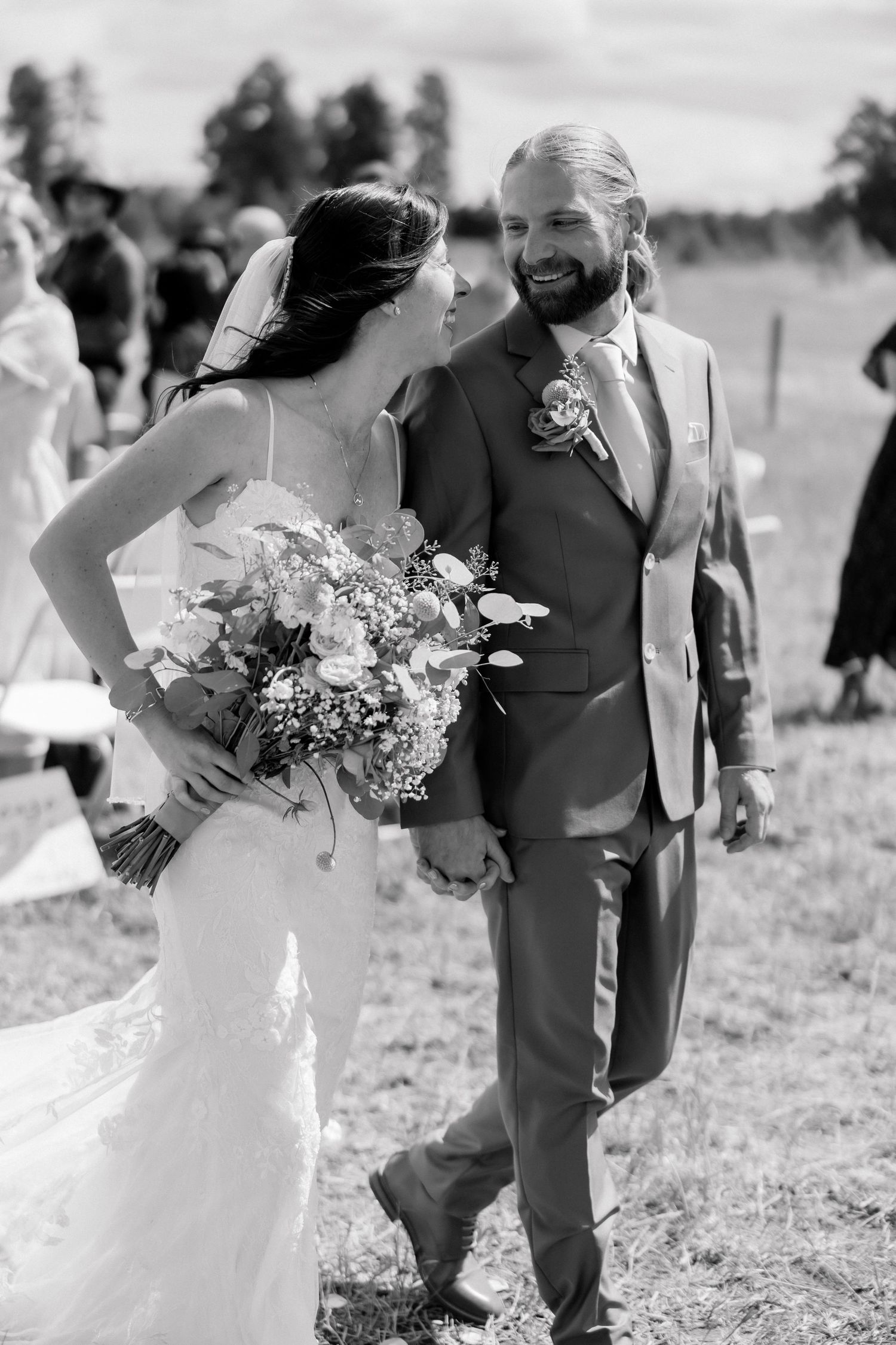 Black and white photo of a smiling bride holding a bouquet and bearded groom in suit walking together after wedding ceremony.