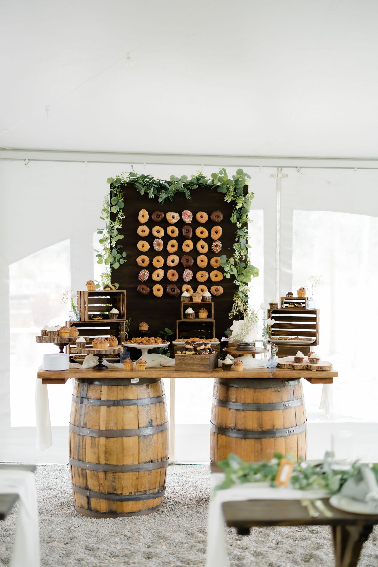 Rustic wedding dessert table with donut wall, wooden barrels, and greenery decor inside a white tent.