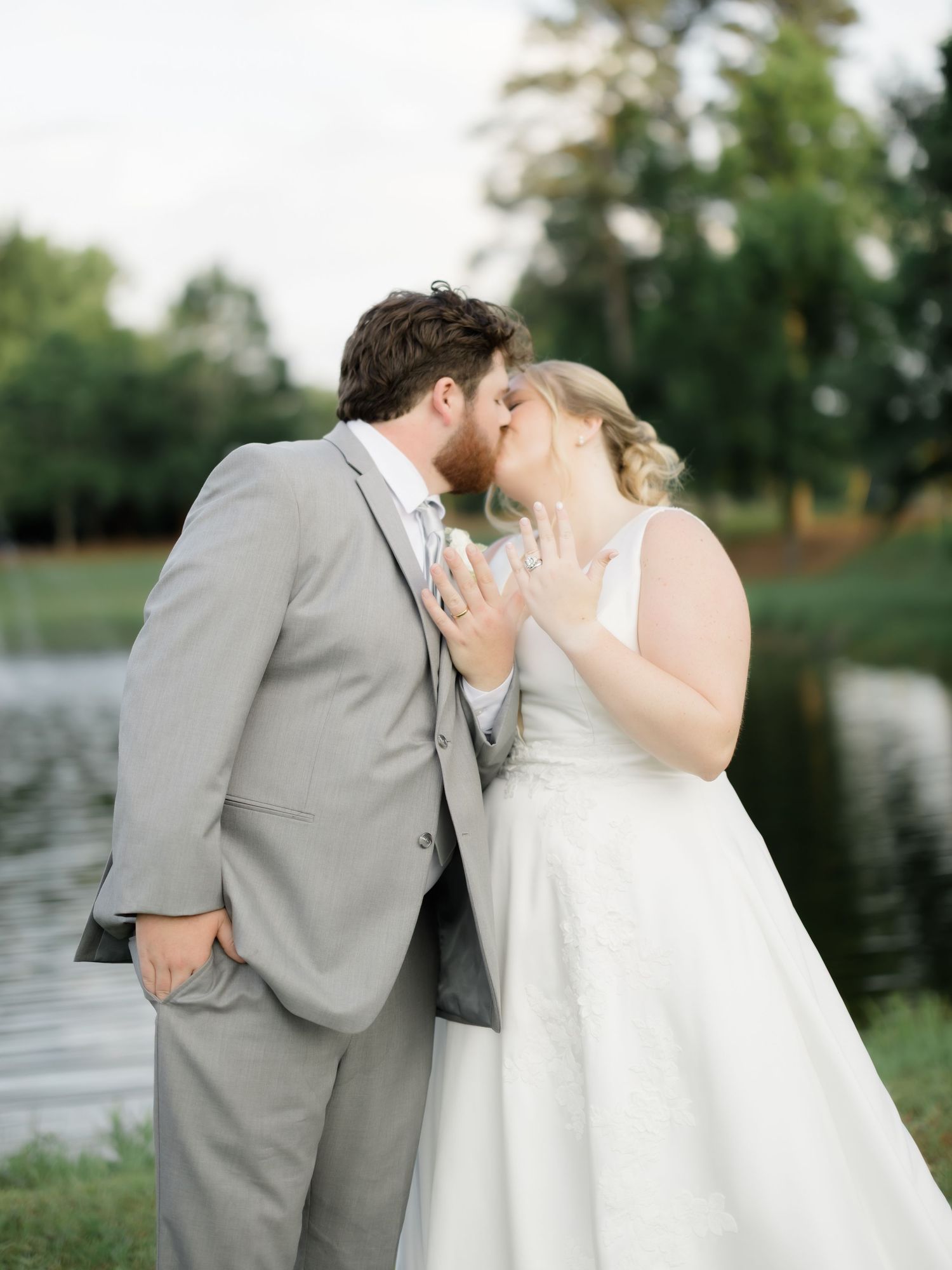 A bride and groom share a romantic kiss outdoors near a pond, with the bride displaying her ring hand.