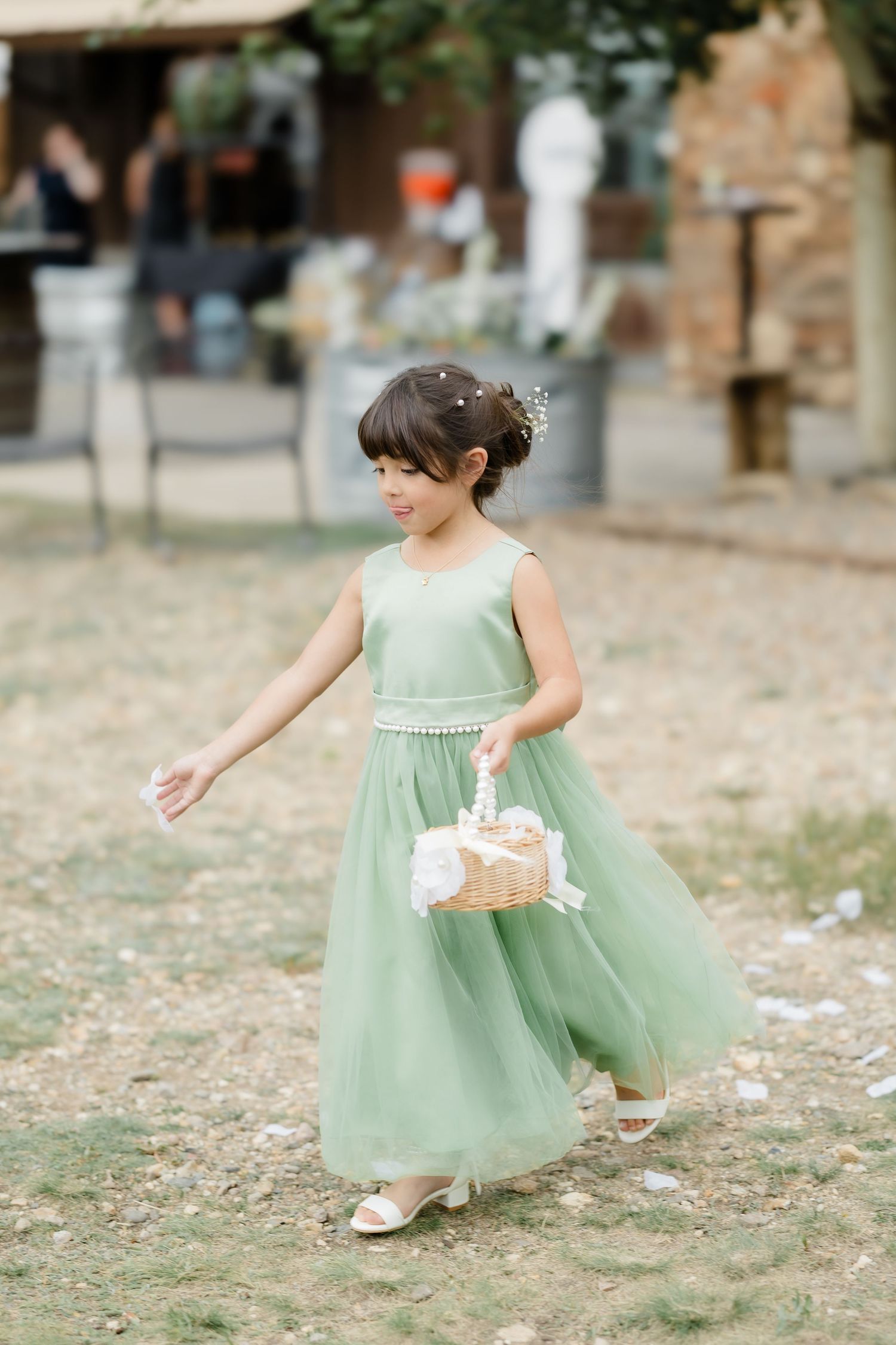 Young flower girl in sage green tulle dress scattering petals at outdoor wedding ceremony.