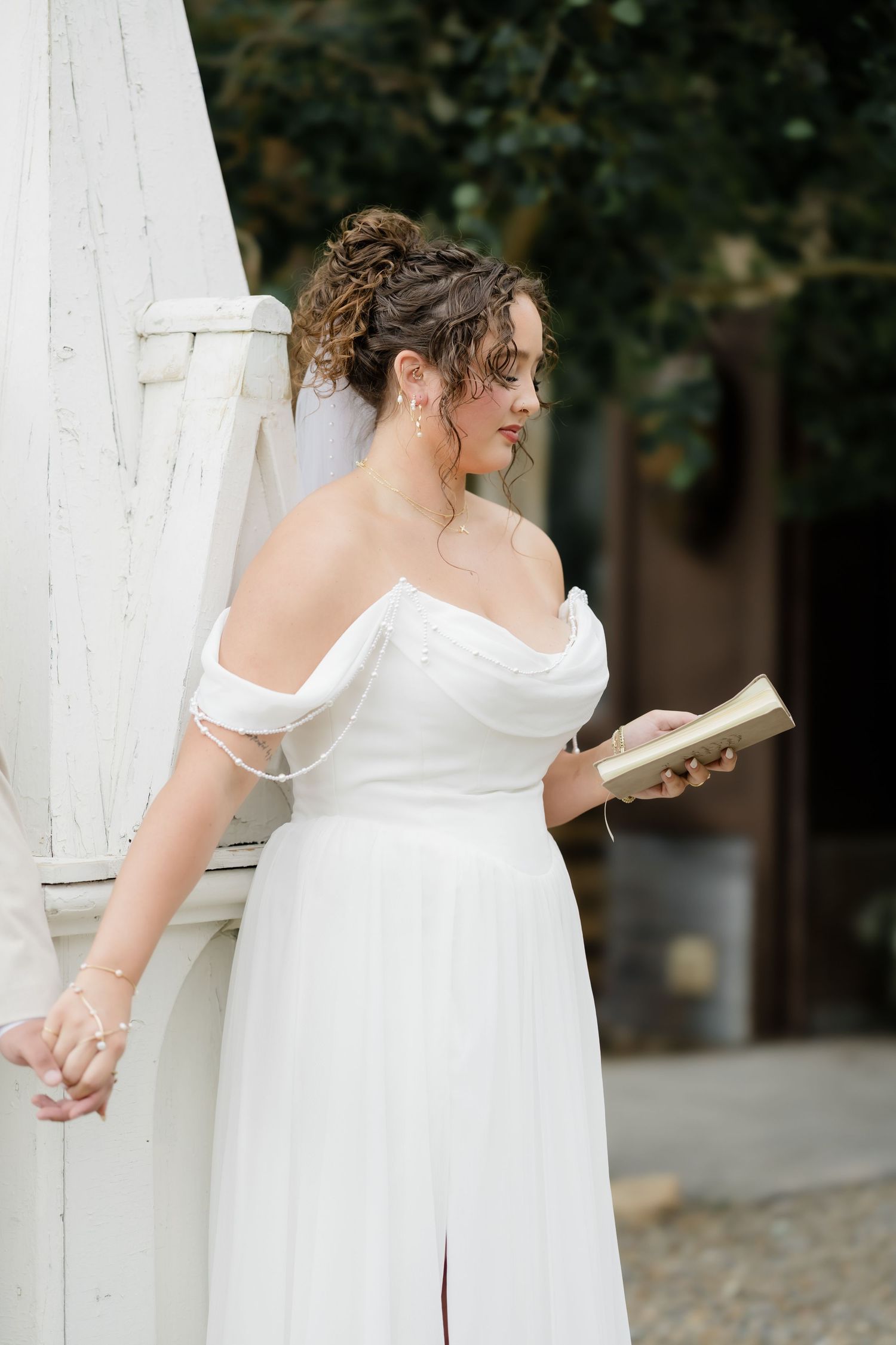 Bride in off-shoulder white gown holding vows book, leaning against white pillar with greenery backdrop.