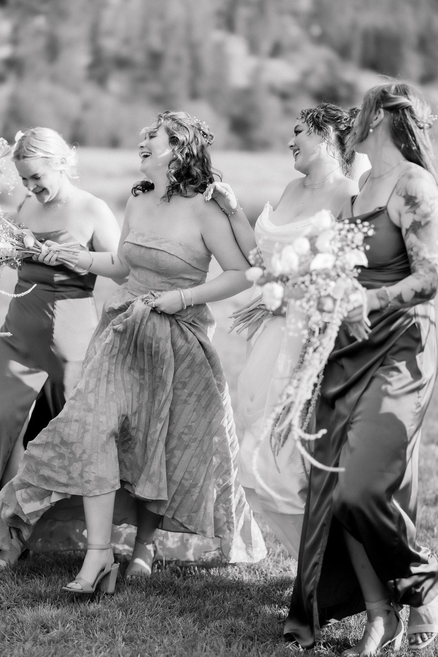 Black and white photo of a bride holding bouquet laughing with bridesmaids walking outdoors at a wedding.