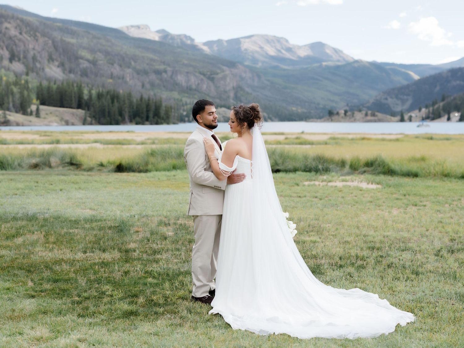 Bride and groom share a romantic moment in a scenic mountain meadow with a lake and rolling hills in the background.