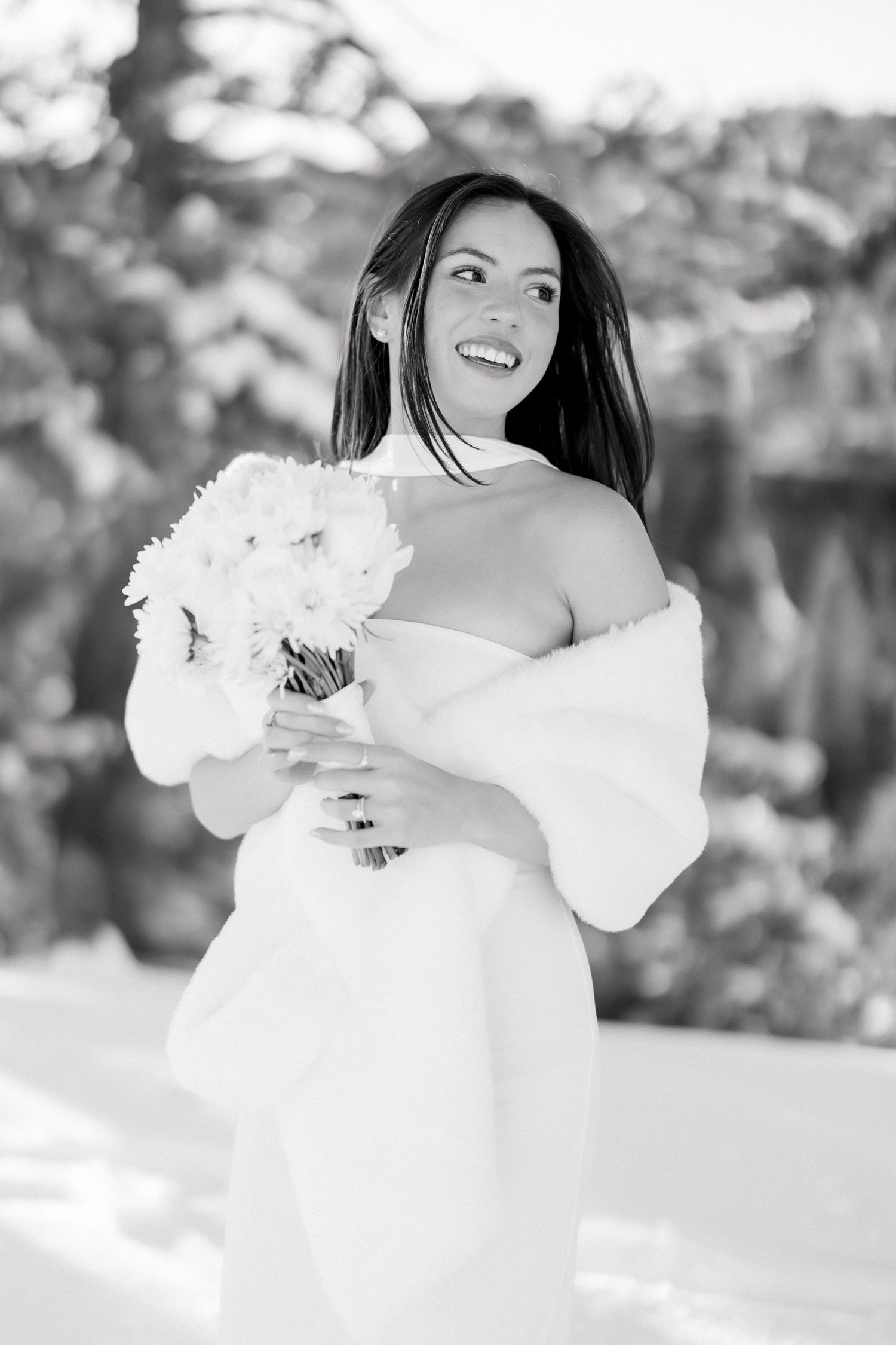 Smiling bride in white strapless gown with fur wrap holds white bouquet outdoors in snowy winter setting.