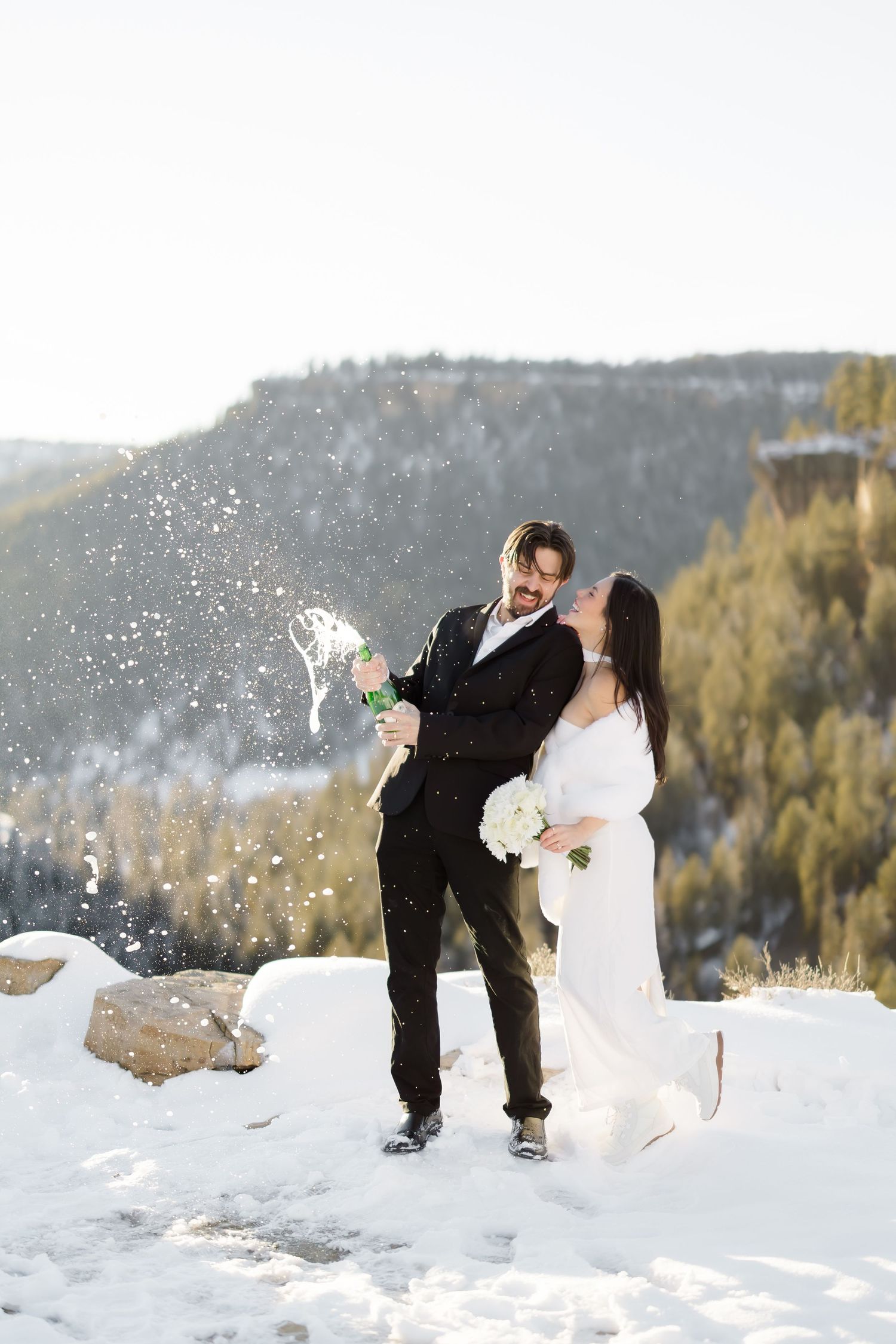 Bride and groom popping champagne in snowy mountain landscape during winter wedding celebration.