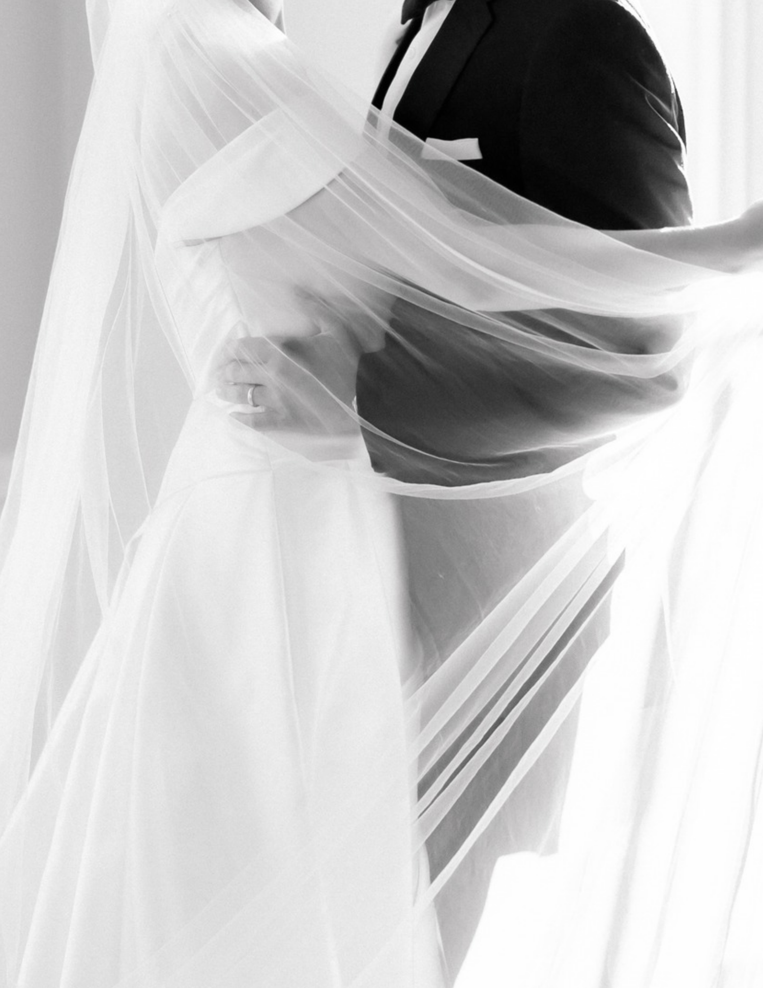 Black and white photo of a bride and groom embracing, her flowing veil billowing around them on their wedding day.