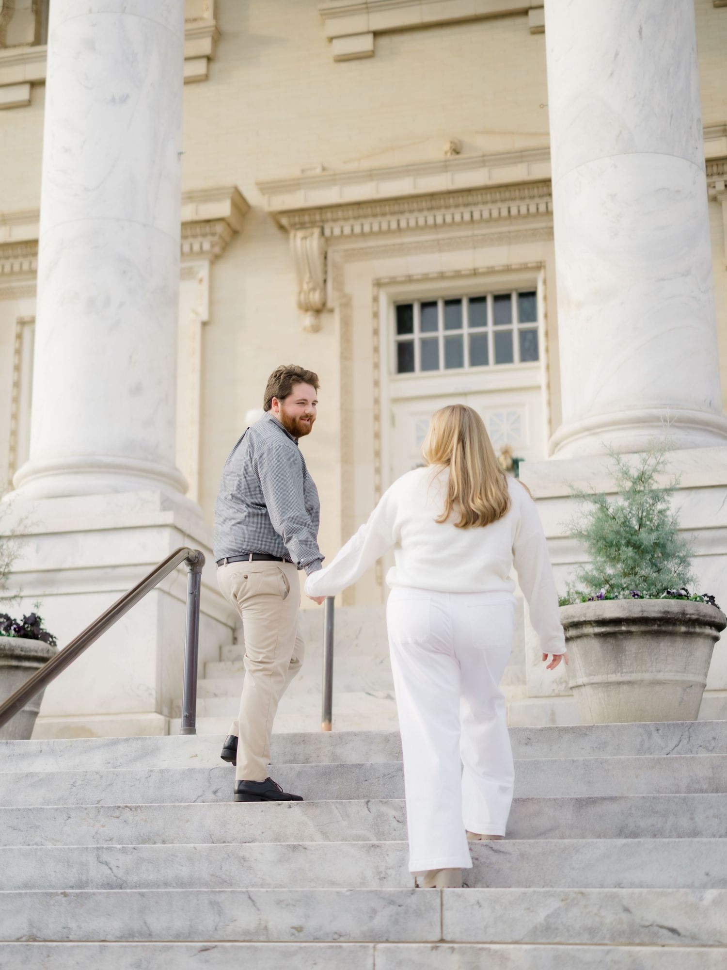 Couple holding hands while climbing marble steps toward a grand classical building entrance.