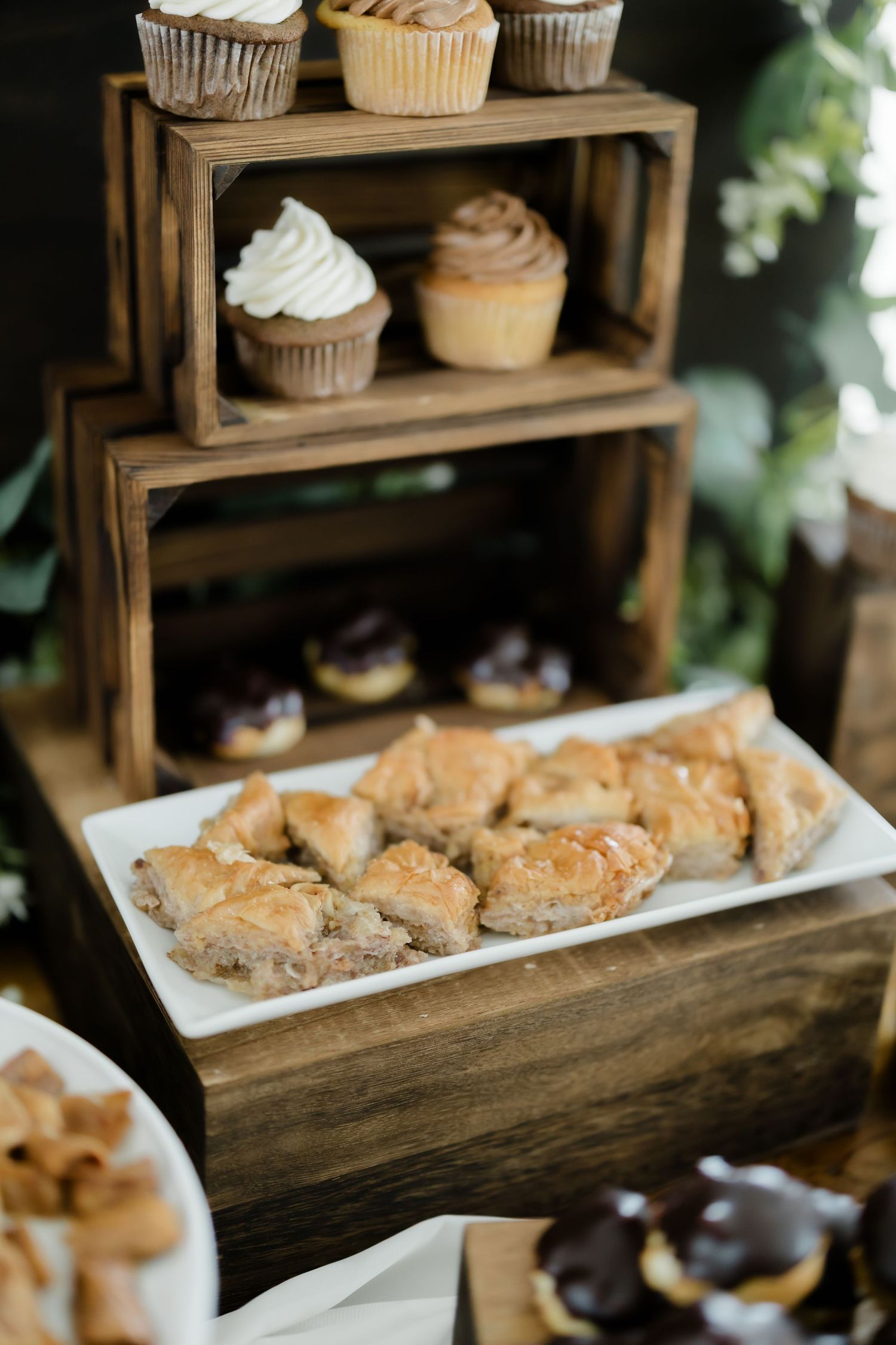 Rustic wooden tiered dessert stand displaying cupcakes and a white plate of golden baklava at a wedding reception.