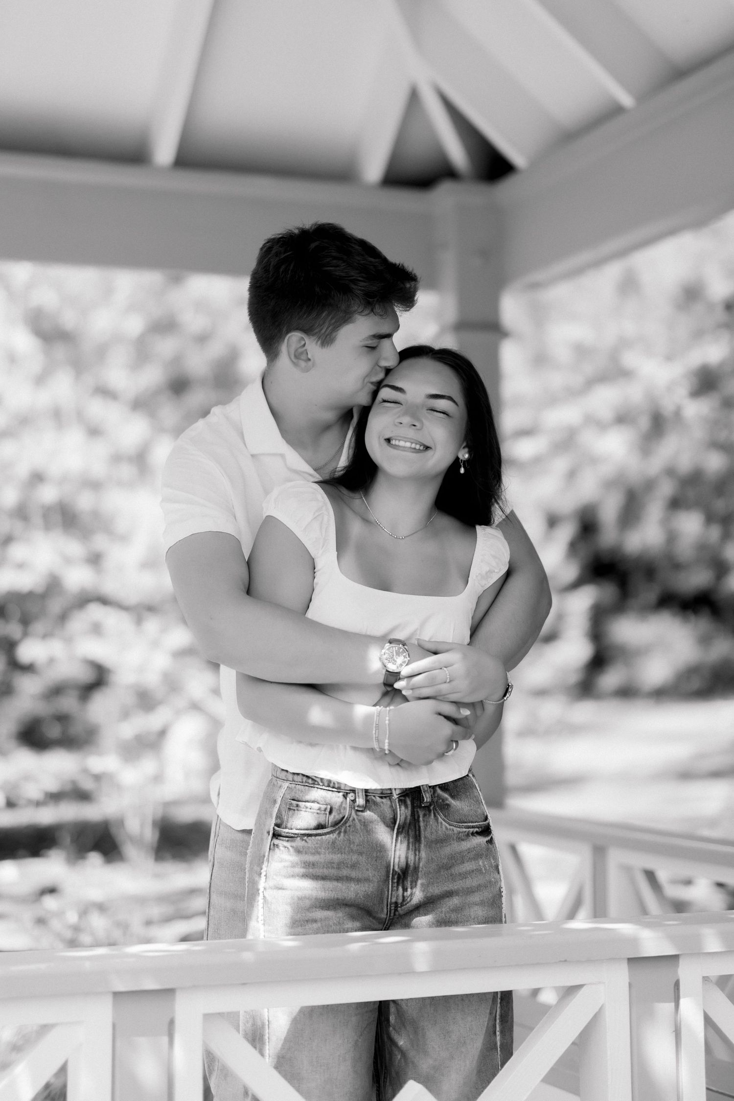 Black and white photo of a young man kissing a smiling woman on the cheek in a gazebo.