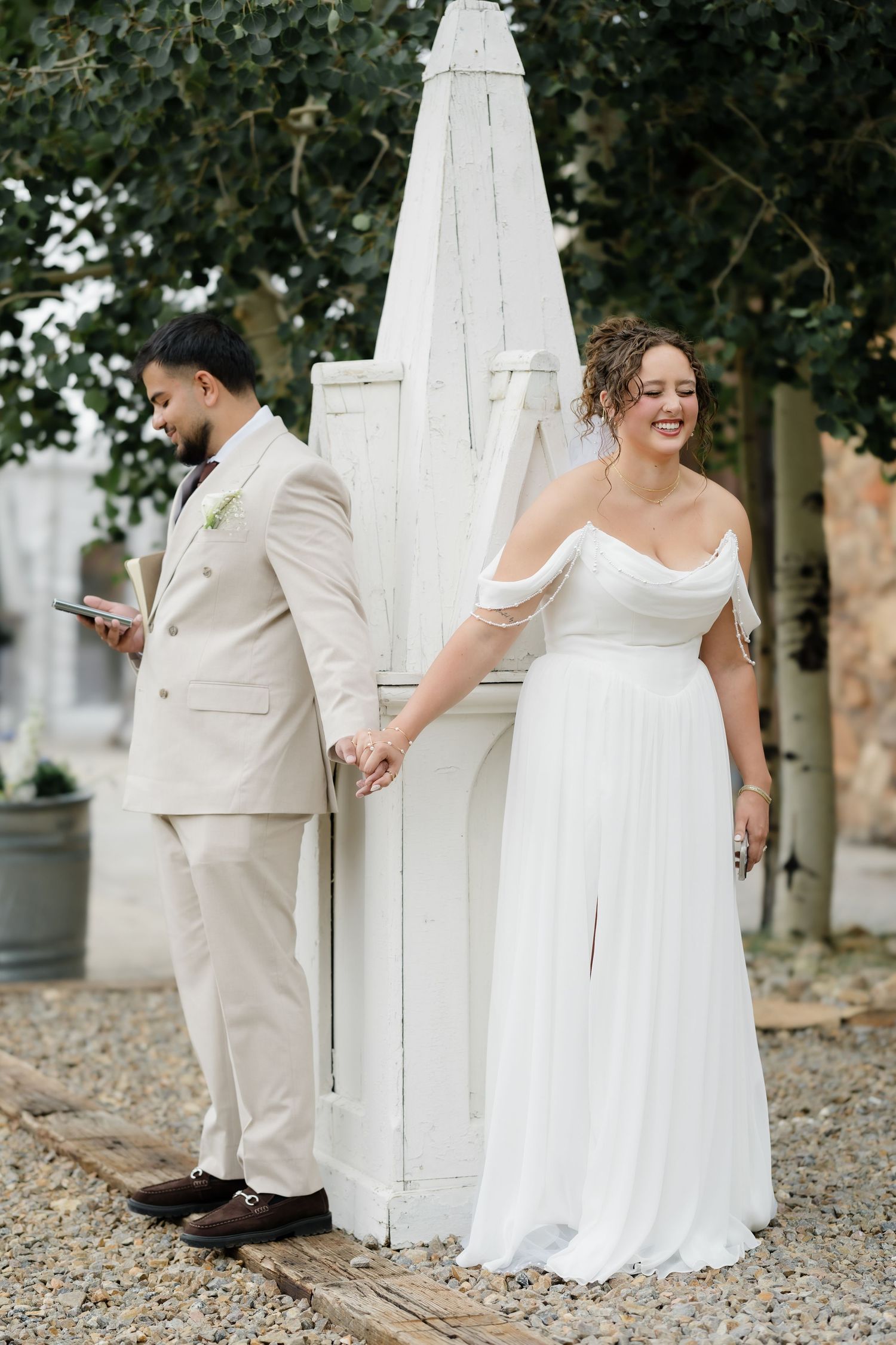 A bride in a white off-shoulder gown holds hands with a groom in a beige suit near a white wooden structure.