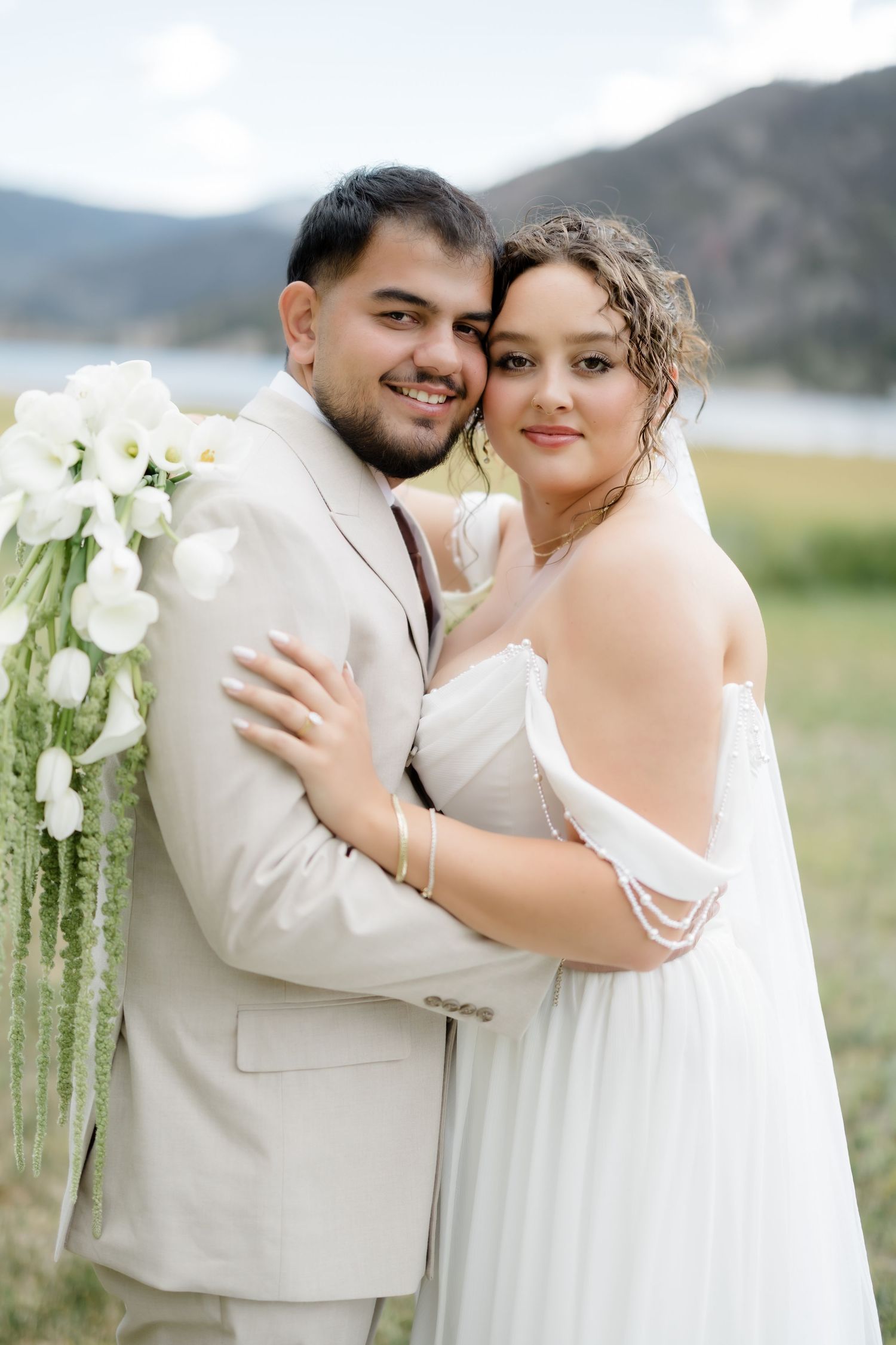 Smiling couple embraces outdoors, bride in white gown with veil, groom in beige suit.