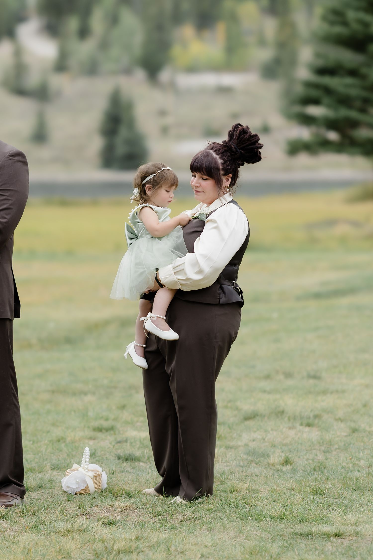 A woman in a brown vest holds a toddler in a green dress at an outdoor wedding ceremony.
