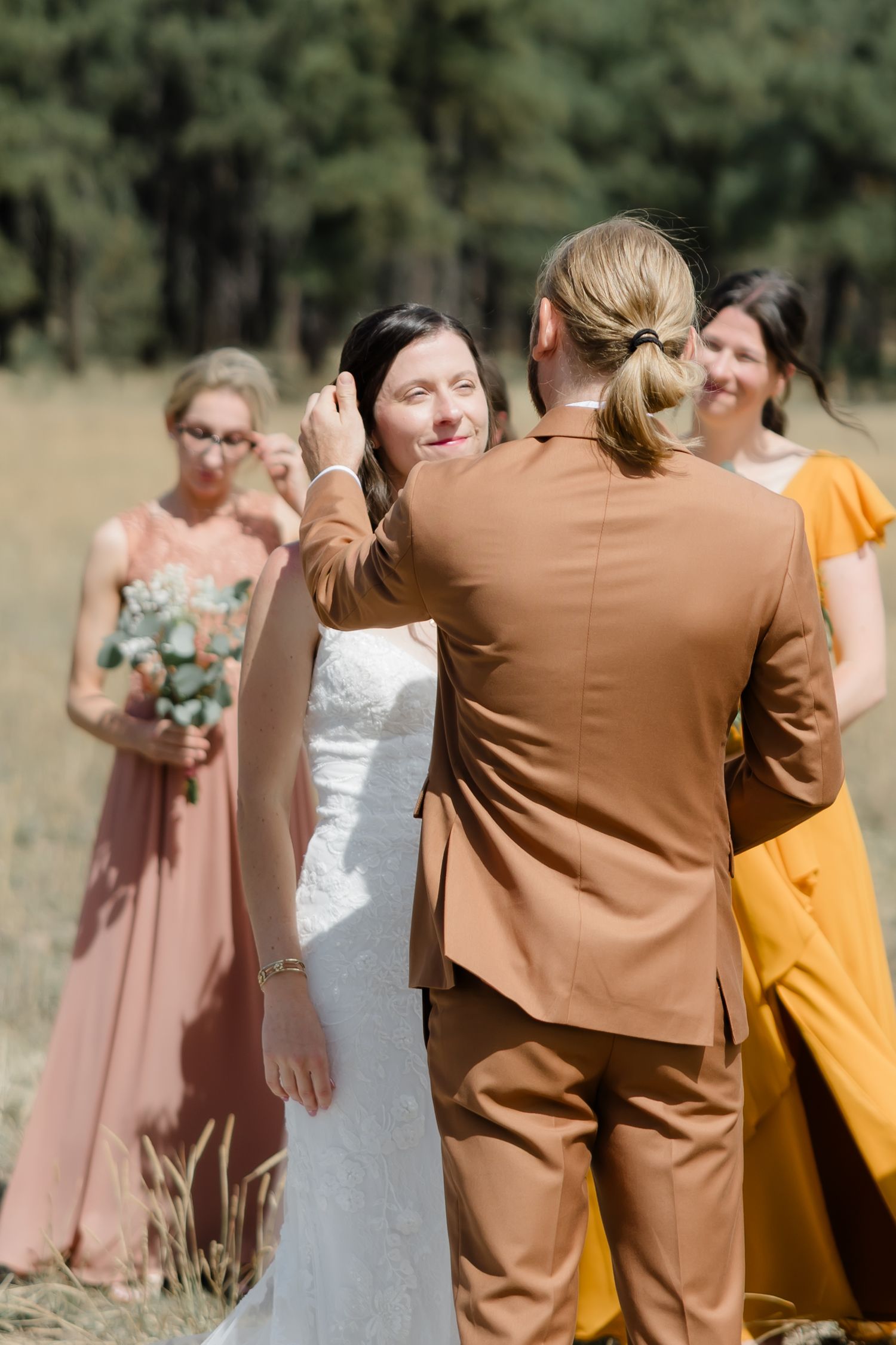 Groom in brown suit tenderly touches brides face during outdoor wedding ceremony with bridesmaids in pink and yellow.