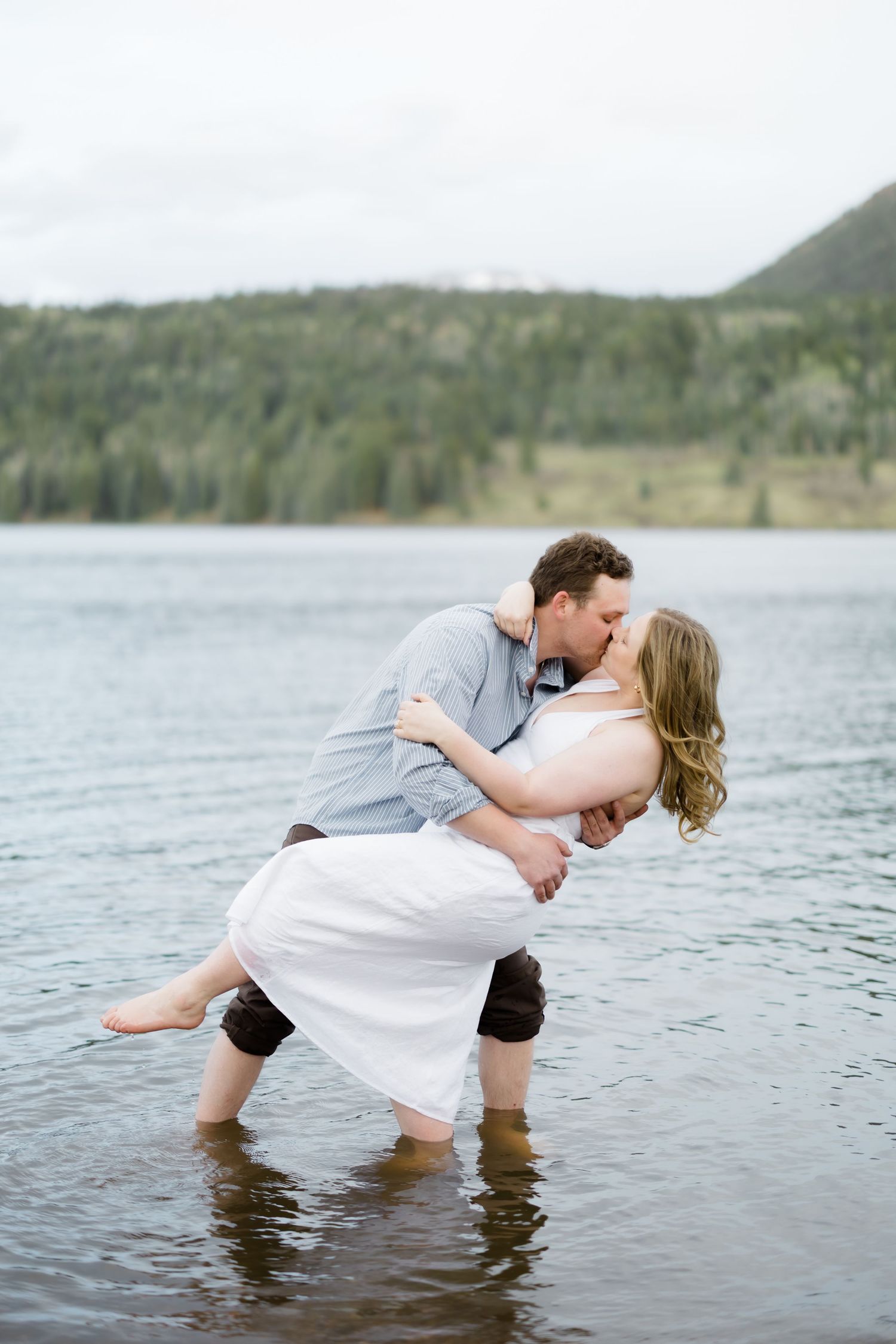 Couple sharing a romantic dip kiss while wading barefoot in a scenic mountain lake together.