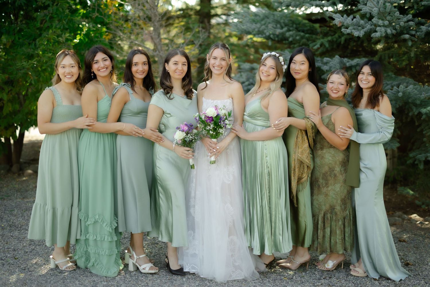 Bride in white gown holds bouquet surrounded by bridesmaids in sage green dresses outdoors.