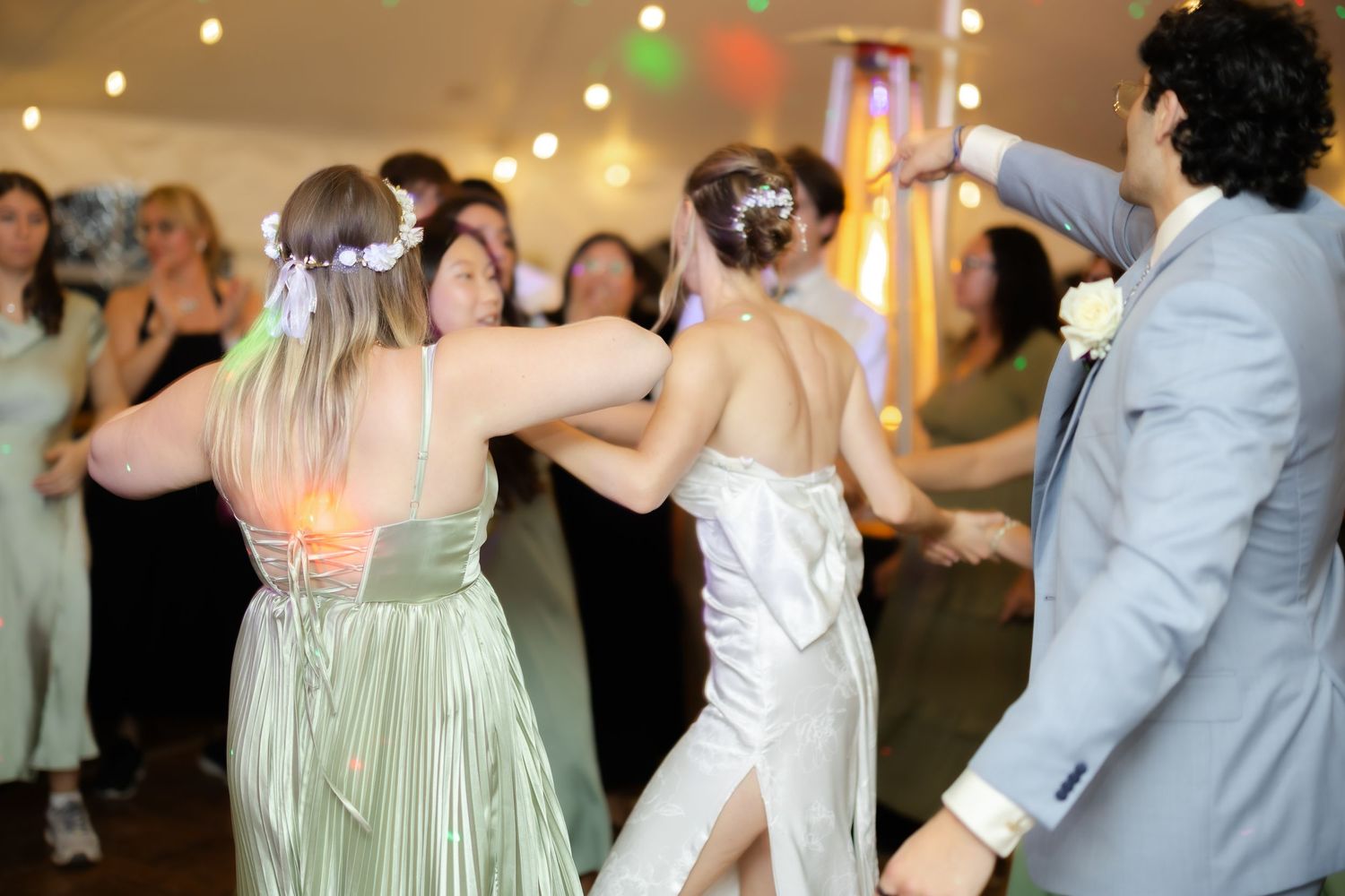 Wedding guests dancing joyfully at a reception, bride in white satin dress with bridesmaids in sage green gowns.