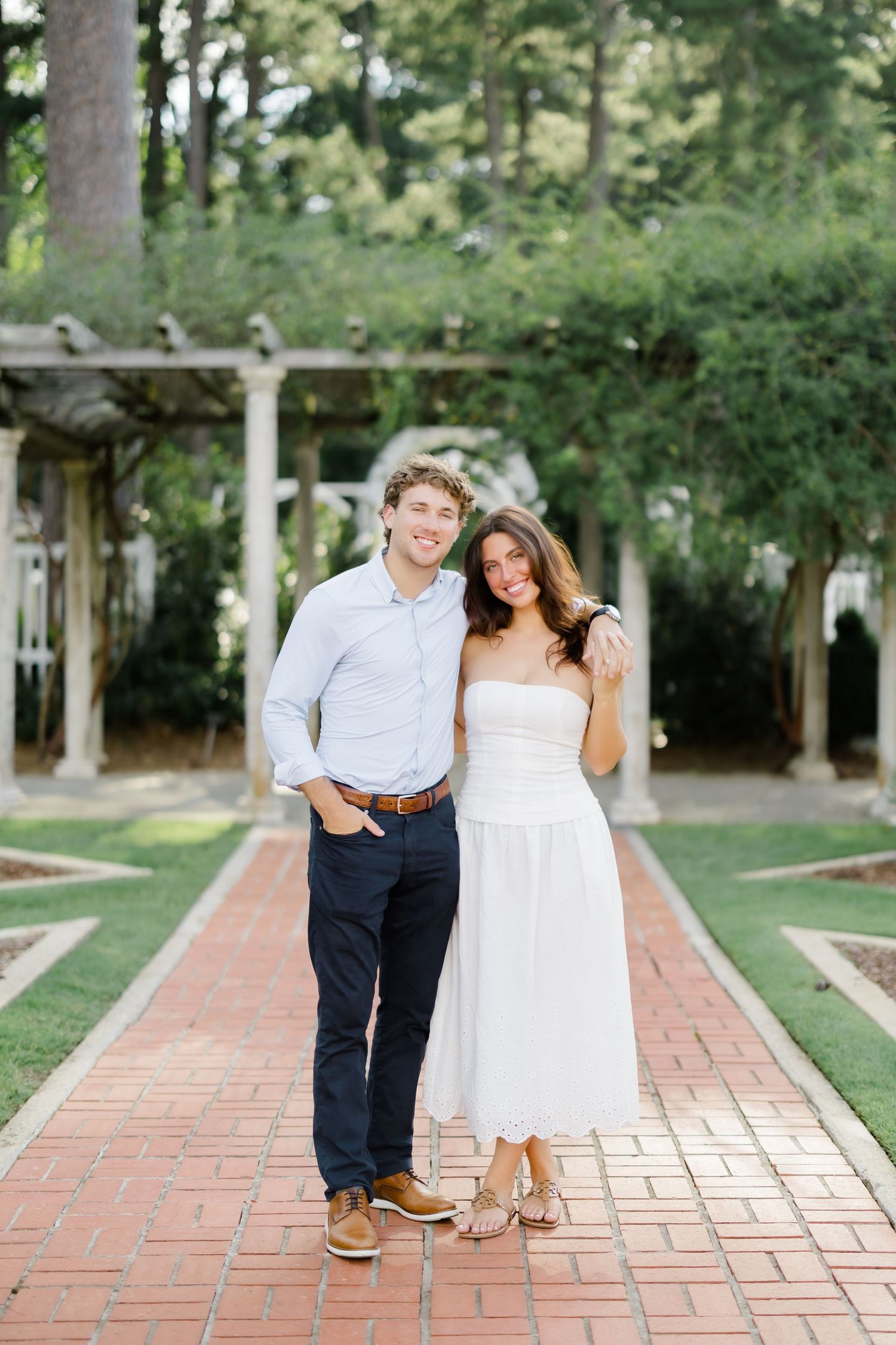 A smiling couple poses on a brick garden path, man in blue shirt and navy pants, woman in white strapless midi dress.